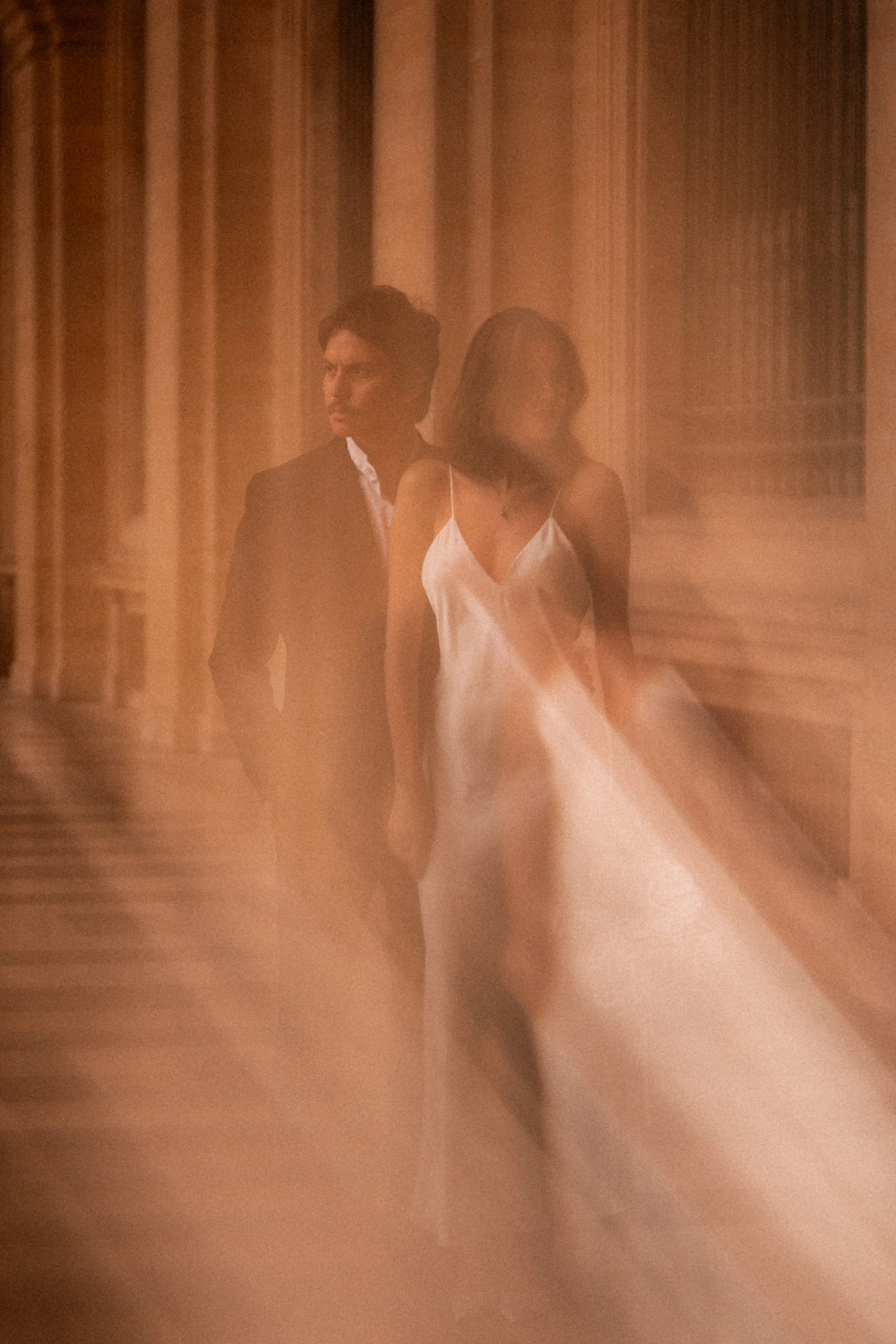 Deux personnes en noir et blanc marchent dans un couloir avec des rideaux en arrière-plan, une femme en robe de soie blanche et un homme en costume