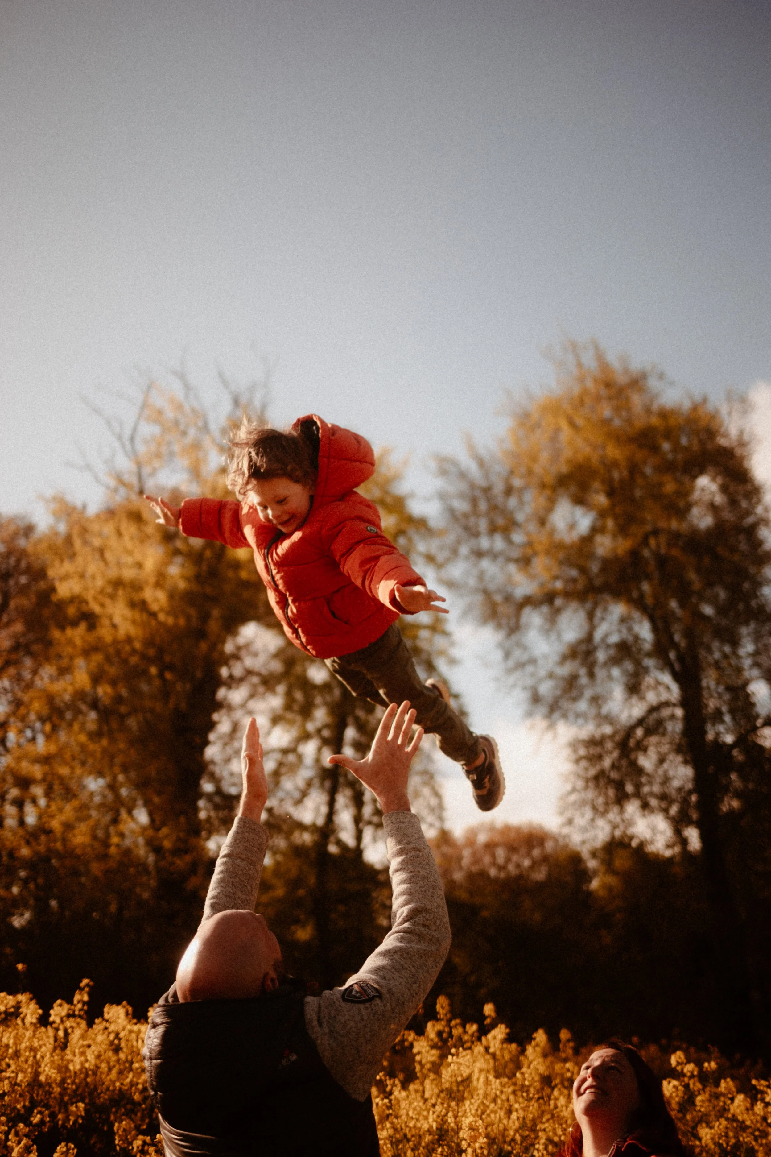 Un enfant en veste rouge est lancé en l'air par un adulte dans un parc en automne, avec des arbres aux feuilles orange et jaune.