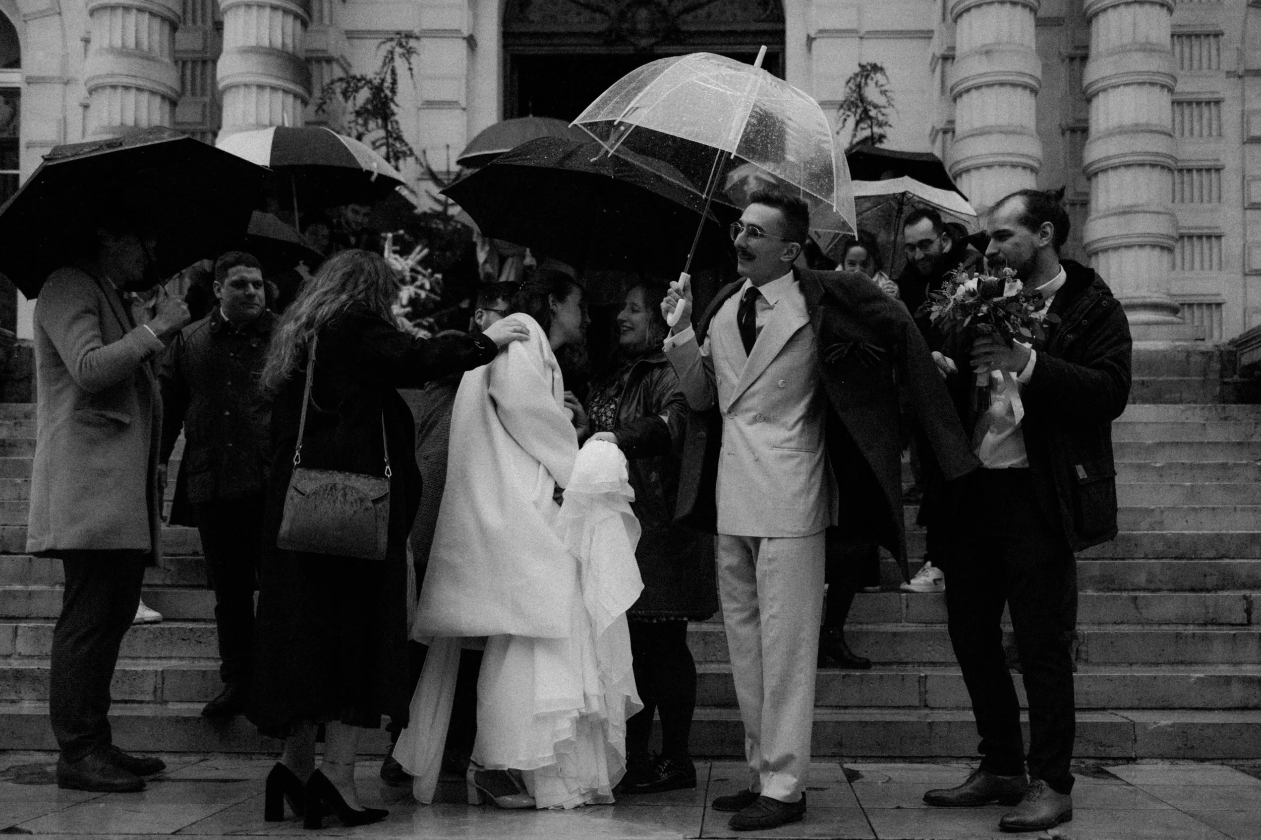 Groupe de personnes en manteaux sous parapluies, en cérémonie en plein air devant un bâtiment historique, ambiance pluvieuse, photographié en noir et blanc.