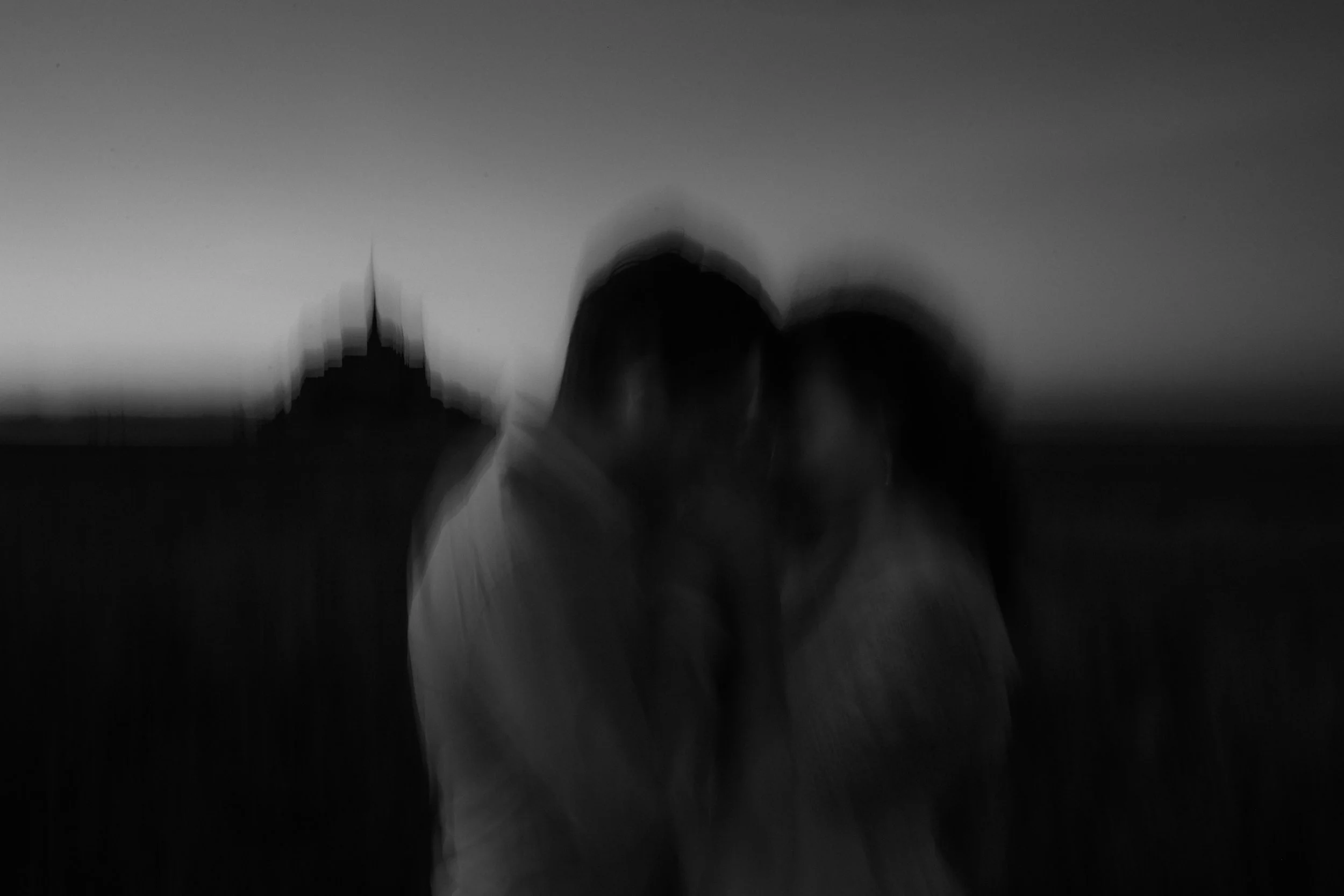 Deux personnes debout près d’un bâtiment en silhouette, en extérieur, à la tombée de la nuit ou en crépuscule, dans une ambiance sombre et floue.