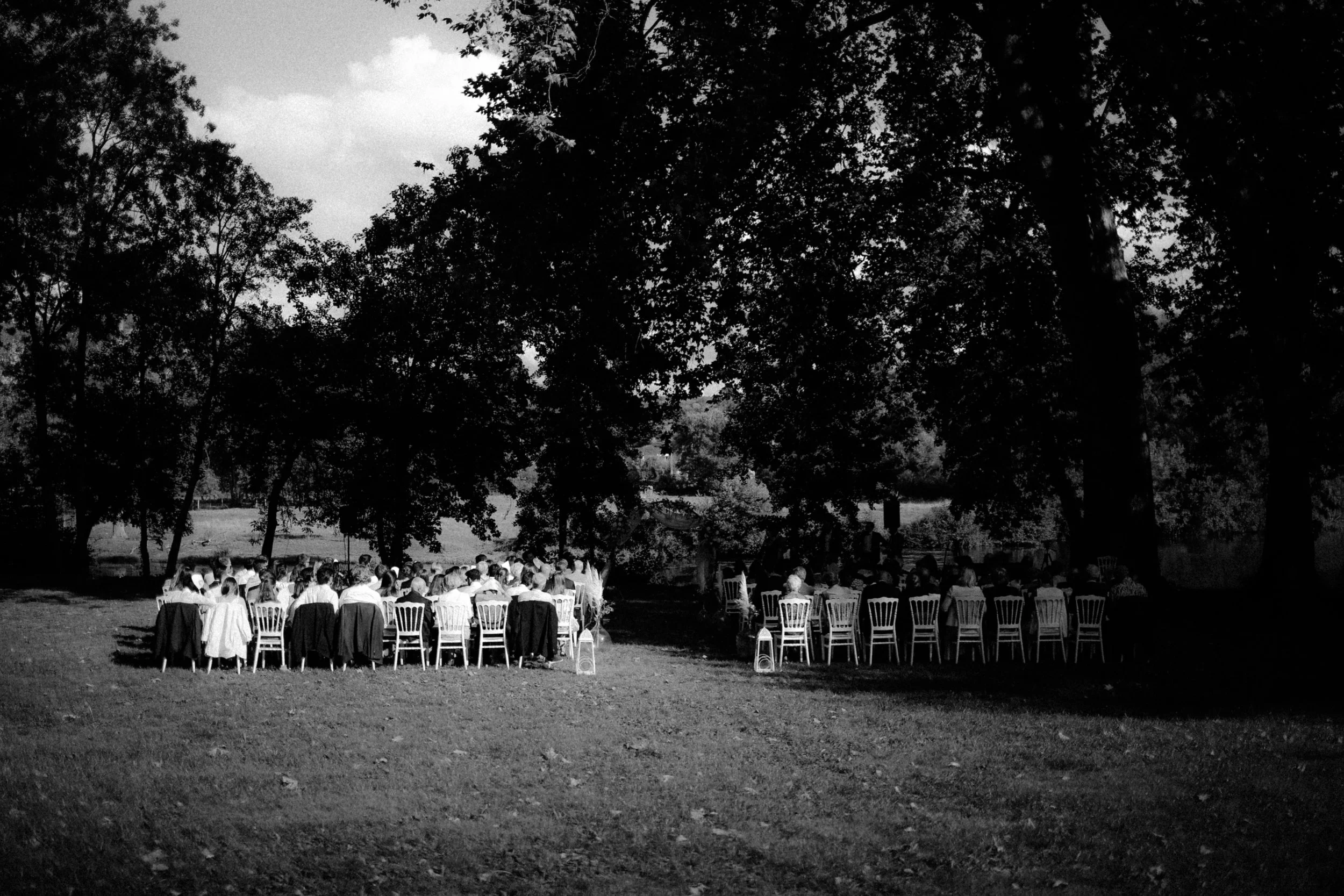 Un groupe de personnes assises à des tables en plein air, entourées d'arbres, lors d'une réunion ou événement en soirée.