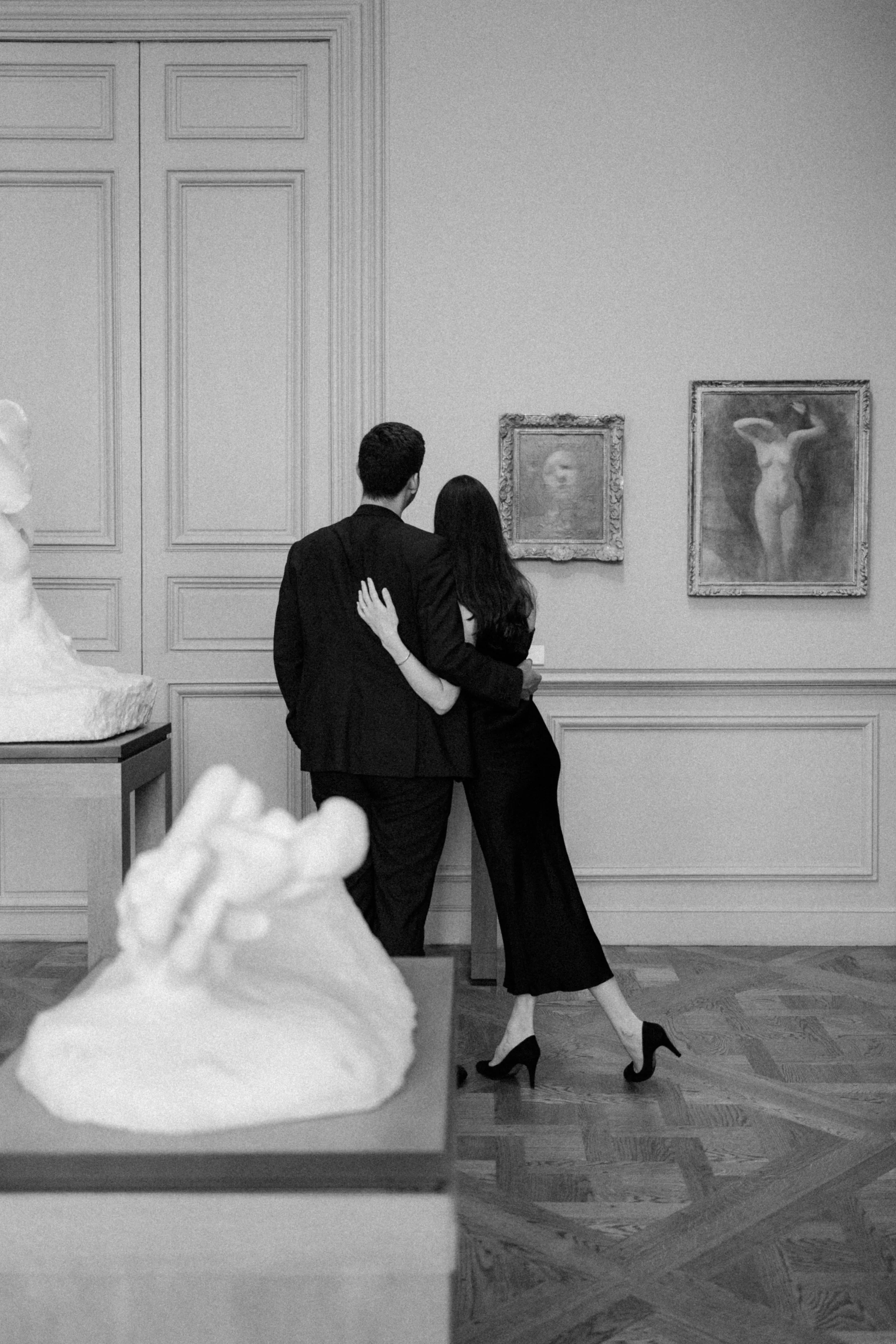 Un couple habillé en noir dans une galerie d'art, regardant des œuvres sur un mur, l'homme avec ses bras autour de la femme, en noir et blanc.