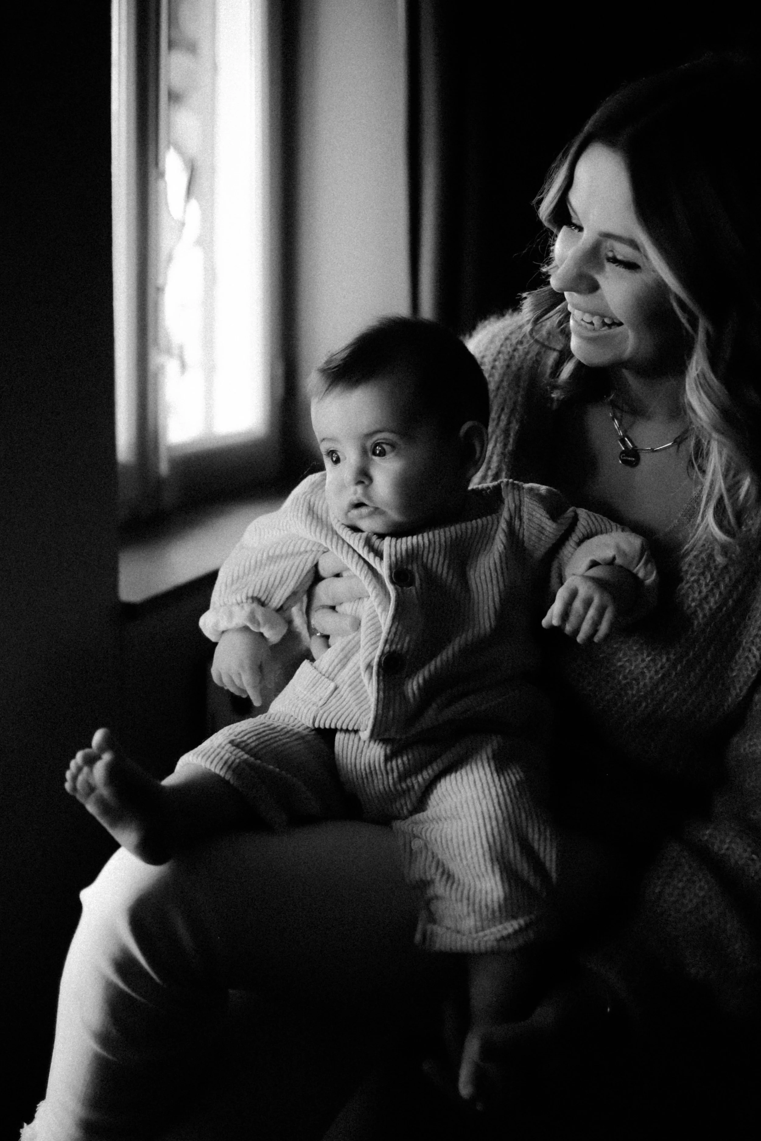 Une femme souriante tient un bébé assis sur ses jambes devant une fenêtre, en noir et blanc.