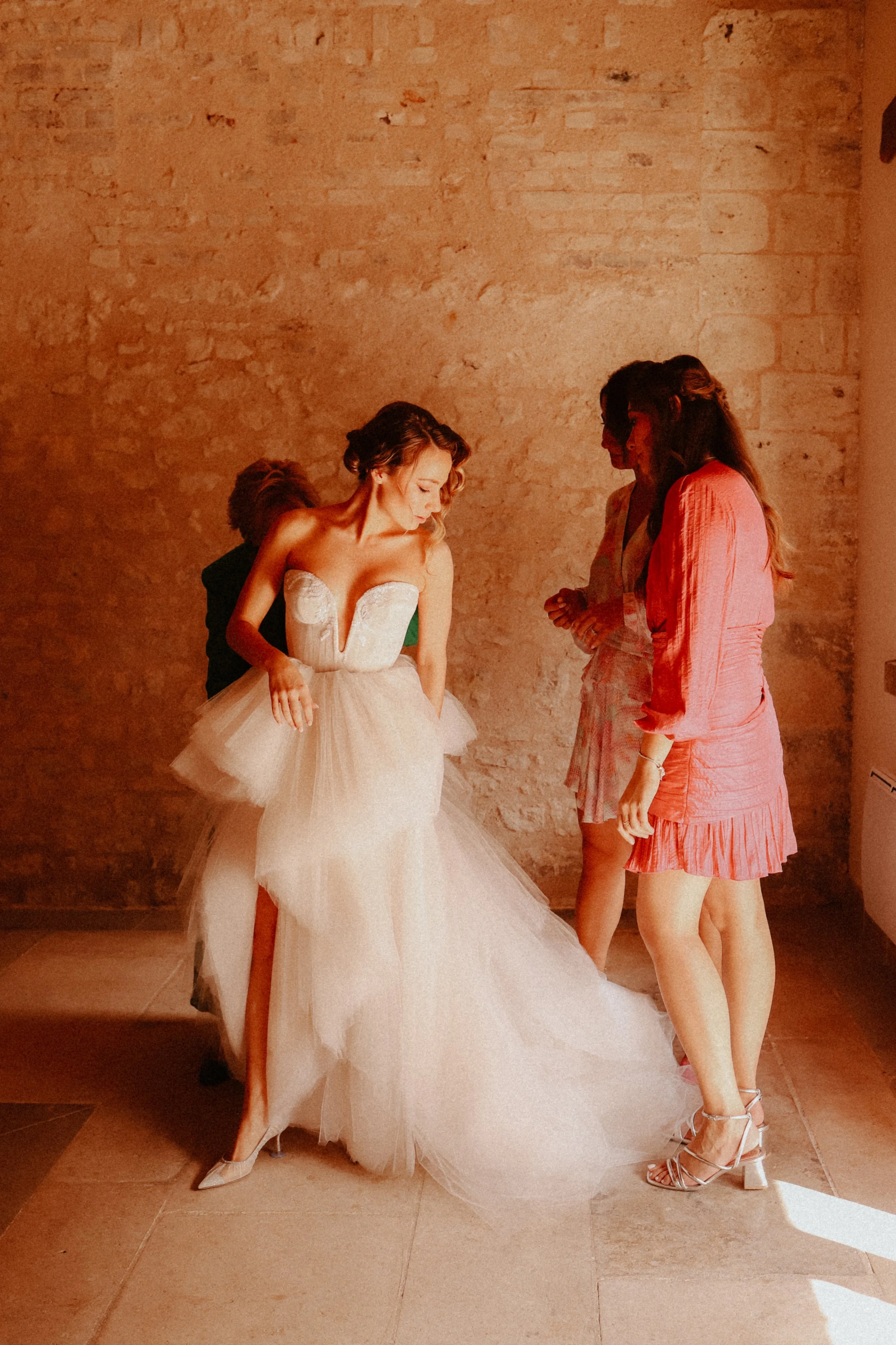Une femme en robe de mariée blanche avec une jupe en tulle, entourée de trois autres femmes en vêtements décontractés. Elles sont dans une pièce avec un mur en pierre visible en arrière-plan.