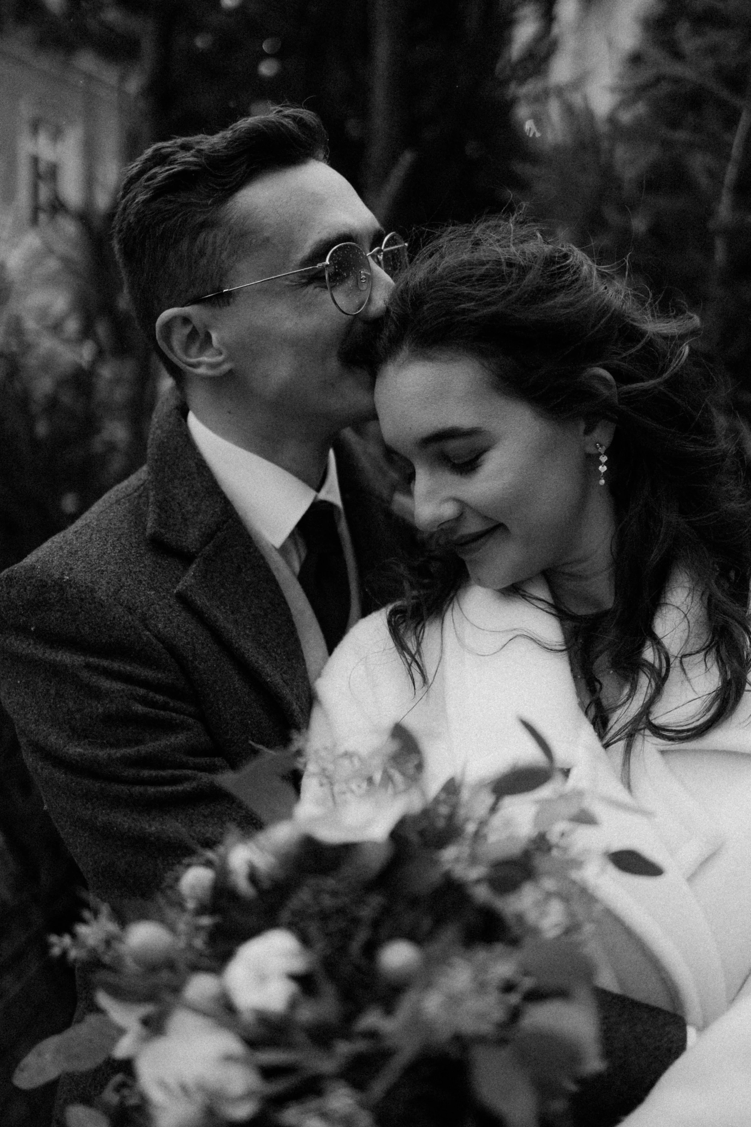 Un homme et une femme en vêtements élégants, partageant un moment intime, avec des fleurs au premier plan, dans un cadre naturel en noir et blanc.