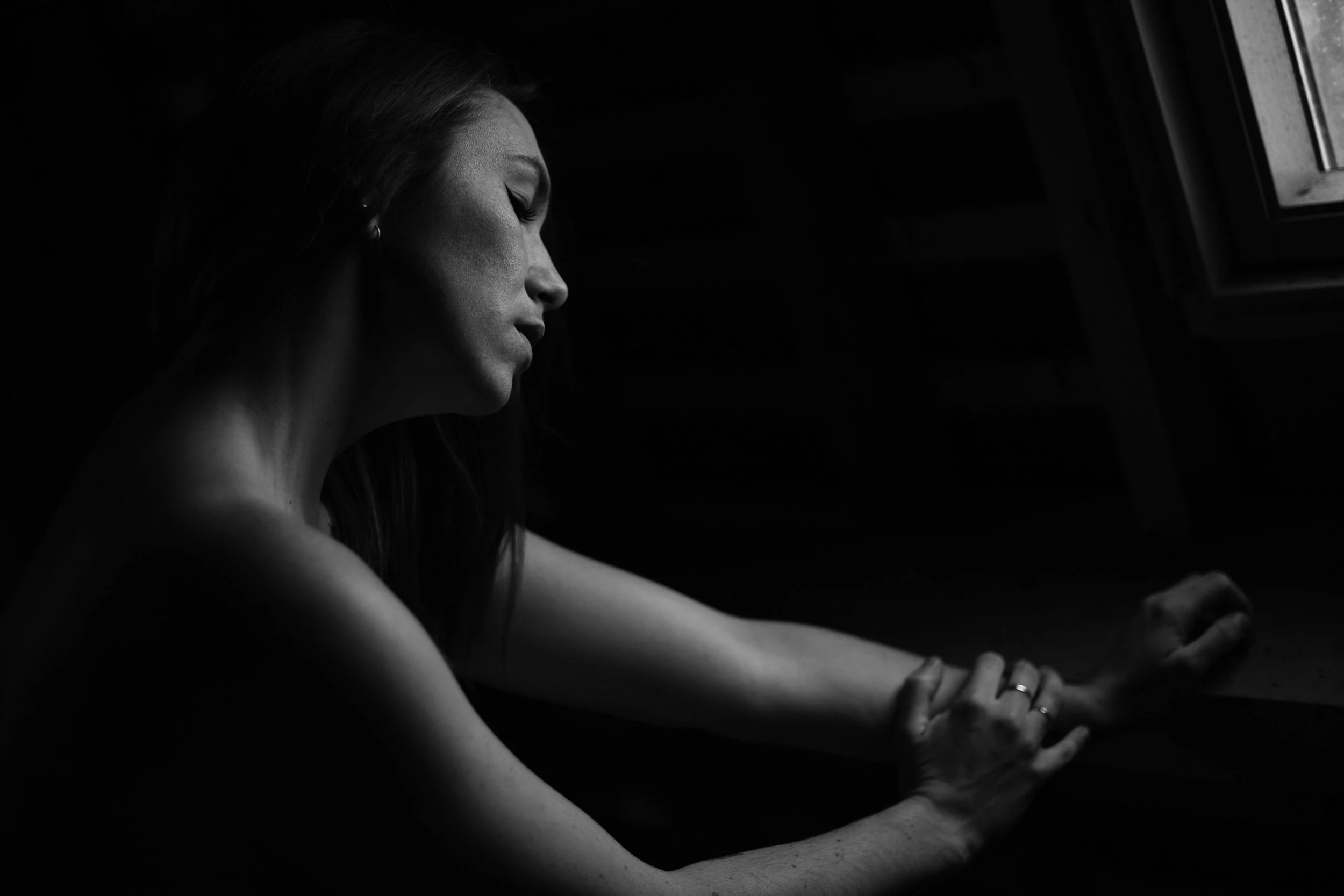 Une femme à la lumière contrastée regarde par une fenêtre, en noir et blanc.