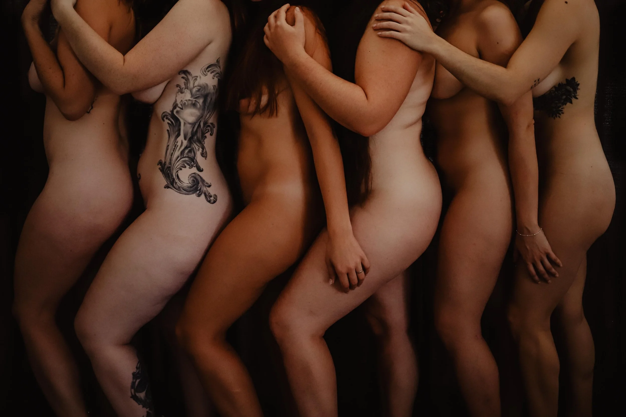 Cinq femmes nues couchant côte à côte, couvrant partiellement leur corps avec leurs bras, certains tatouages visibles sur leurs côtés et bras, pose artistique, fond noir.