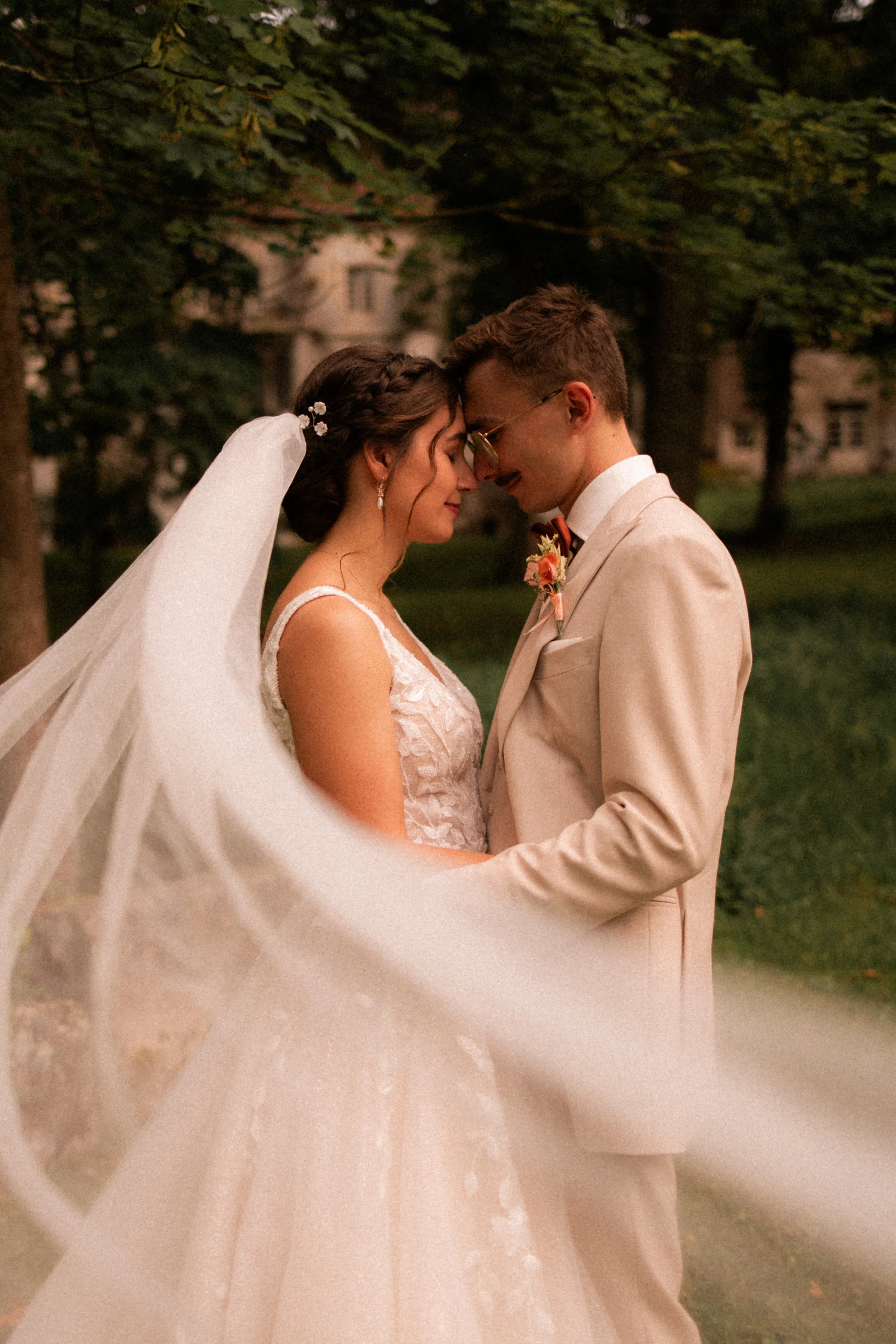 Un couple de mariés se tient face à face, les yeux fermés, dans un parc verdoyant, avec des arbres en arrière-plan.