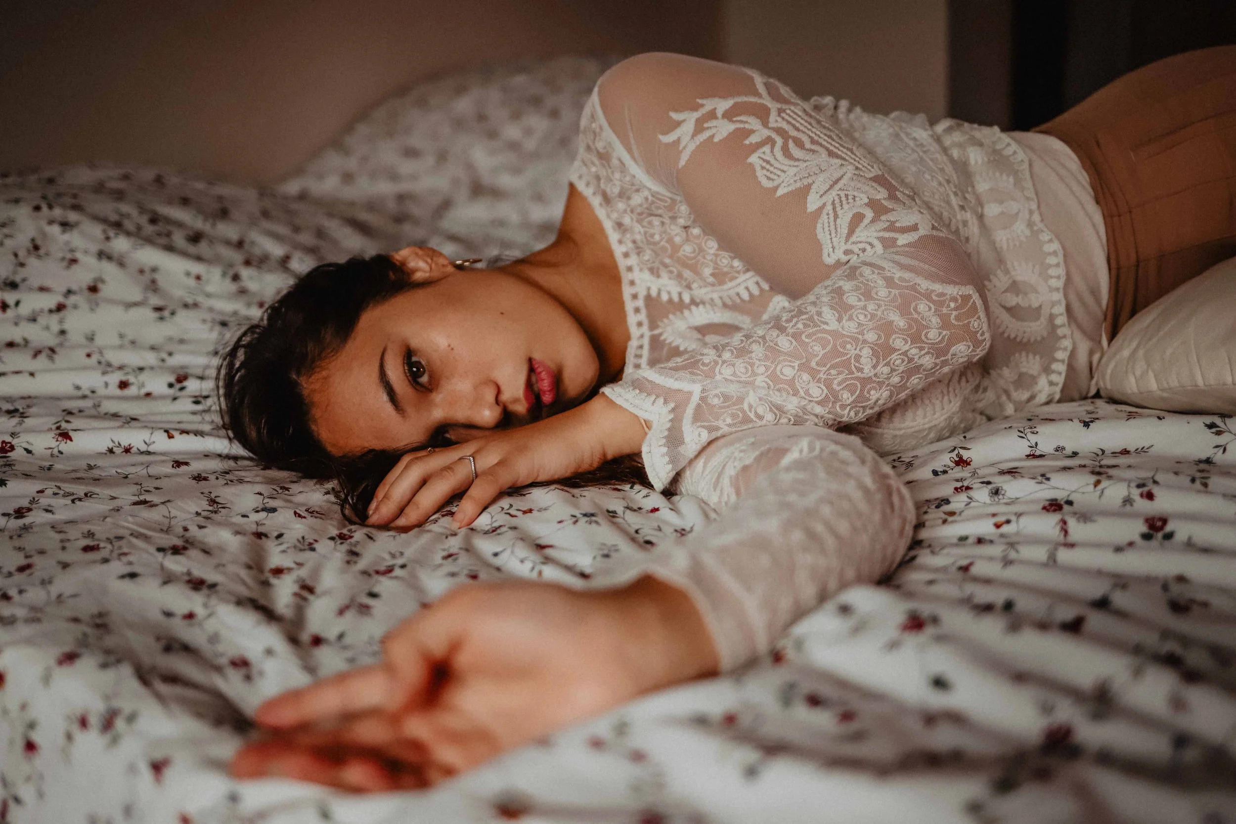 Une femme allongée sur un lit, portant un haut en dentelle blanche et des pantalons marron, regarde l'objectif avec une expression calme, dans une pièce tamisée.