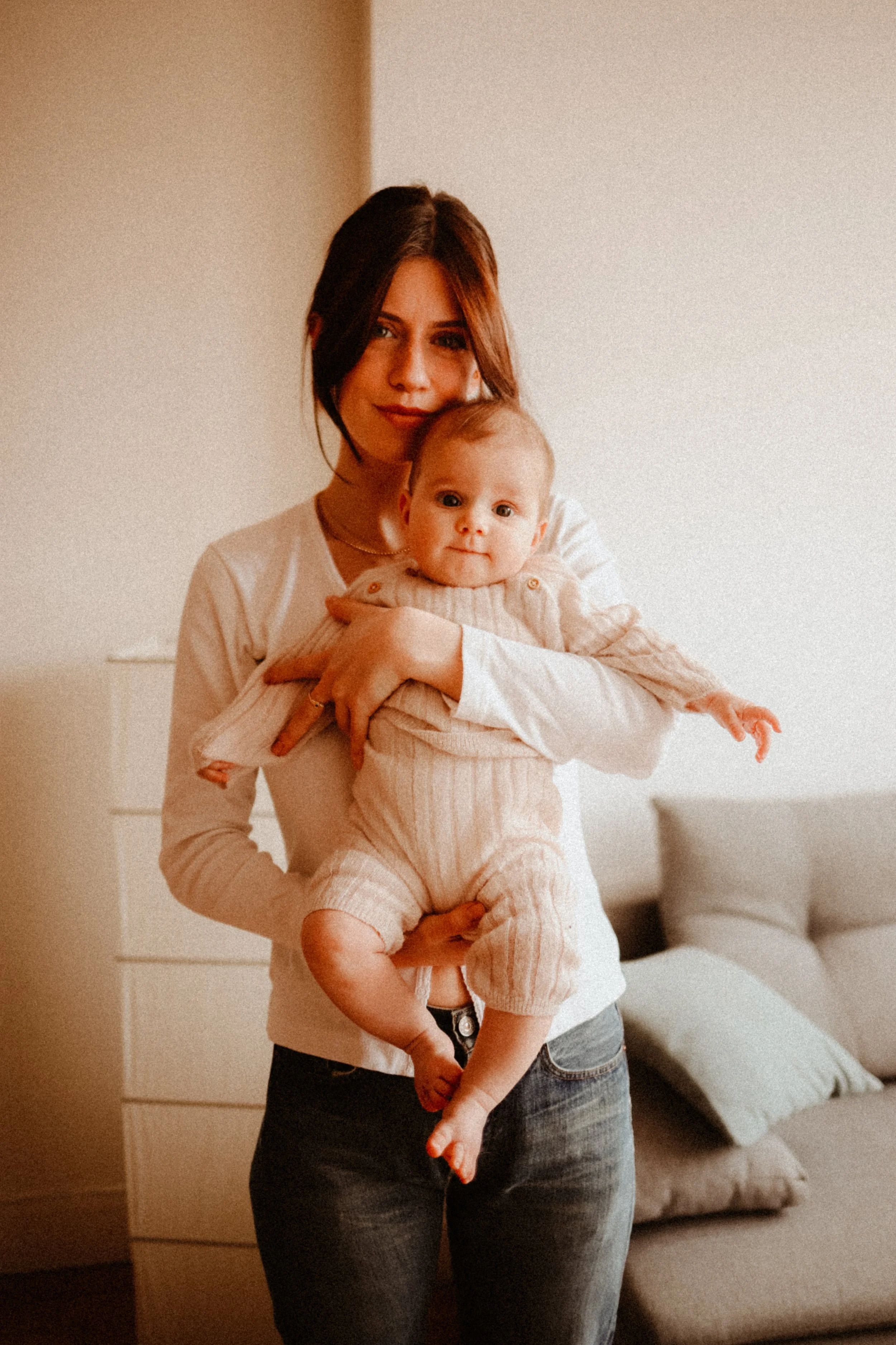 Une femme jeune tenant un bébé dans un salon intérieur, tous deux souriant, avec un canapé en arrière-plan.