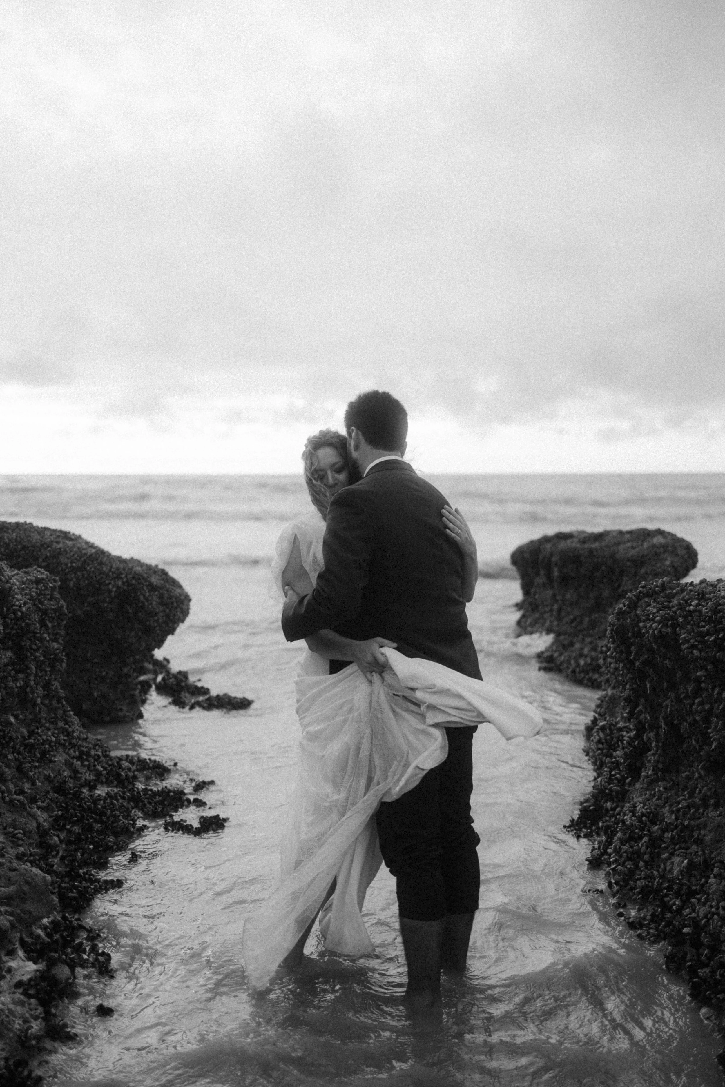 Un couple en vêtements de mariage dans la mer, l'homme tenant la femme dans ses bras, paysage au bord de l'océan en noir et blanc.
