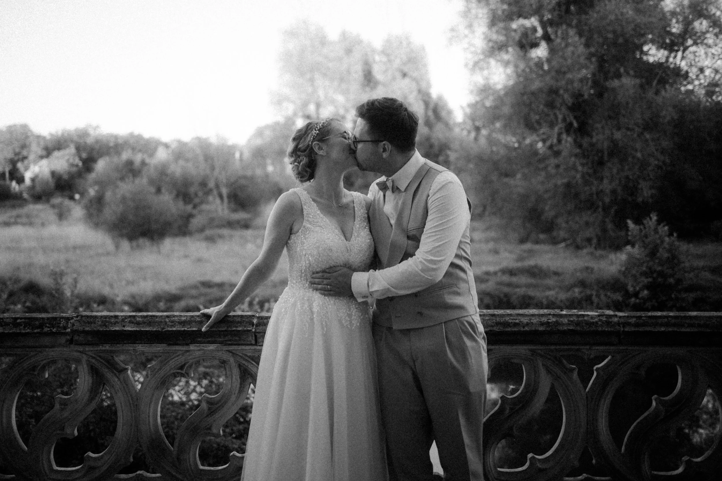 Un couple de mariés s'embrassant sur un pont en fer forgé, en plein air, avec des arbres en arrière-plan, en noir et blanc.