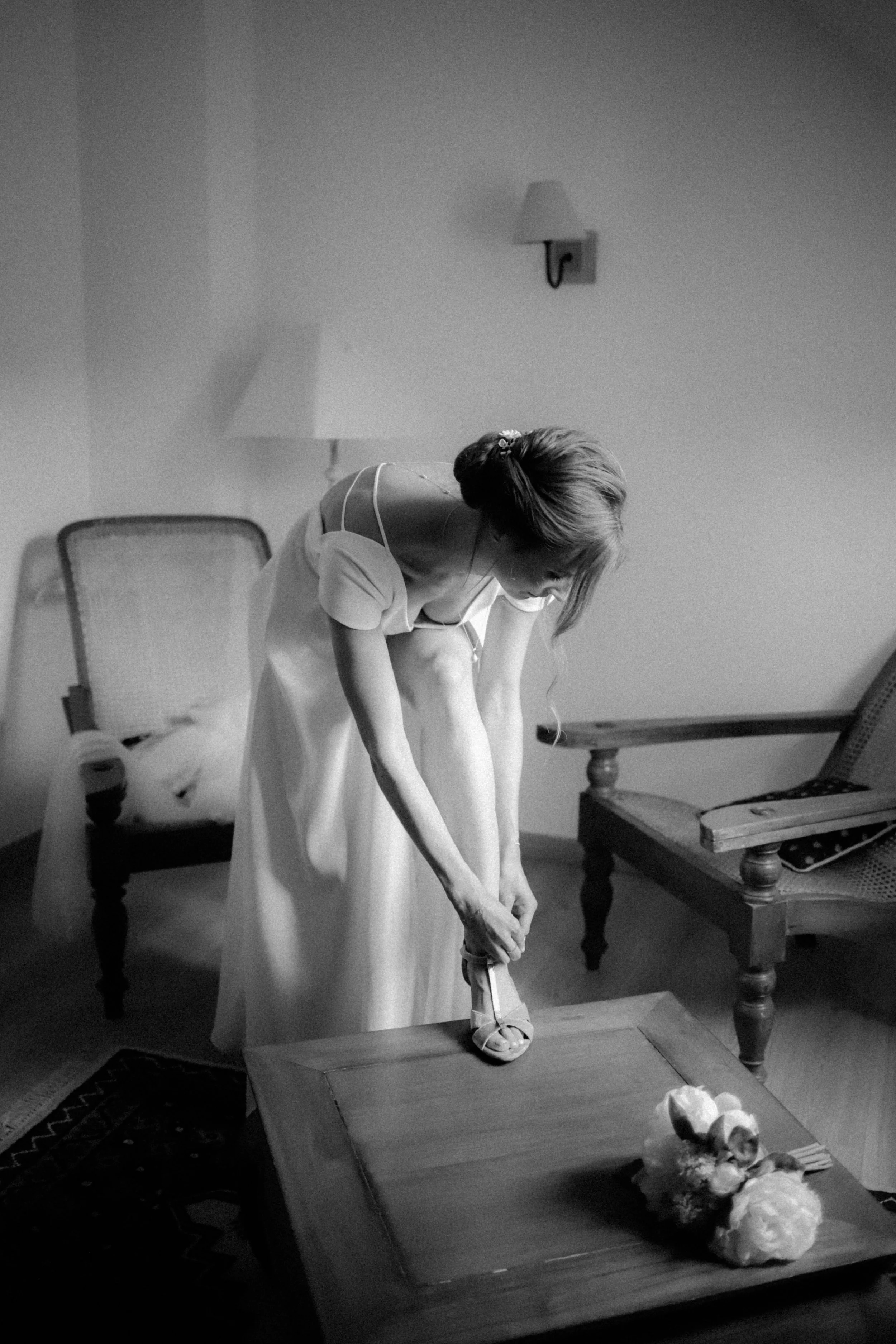 Une femme en robe claire ajuste ses chaussures dans une pièce, avec un fauteuil et une table, en noir et blanc.