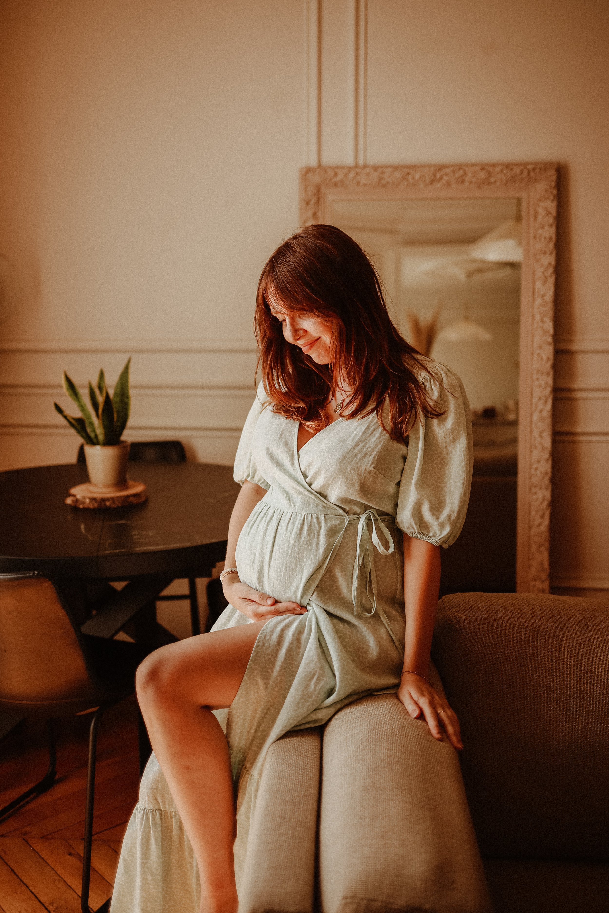 Une femme enceinte assise sur le bord d'un canapé dans une pièce chaleureuse, portant une robe claire avec des manches bouffantes, regardant son ventre avec un sourire doux.