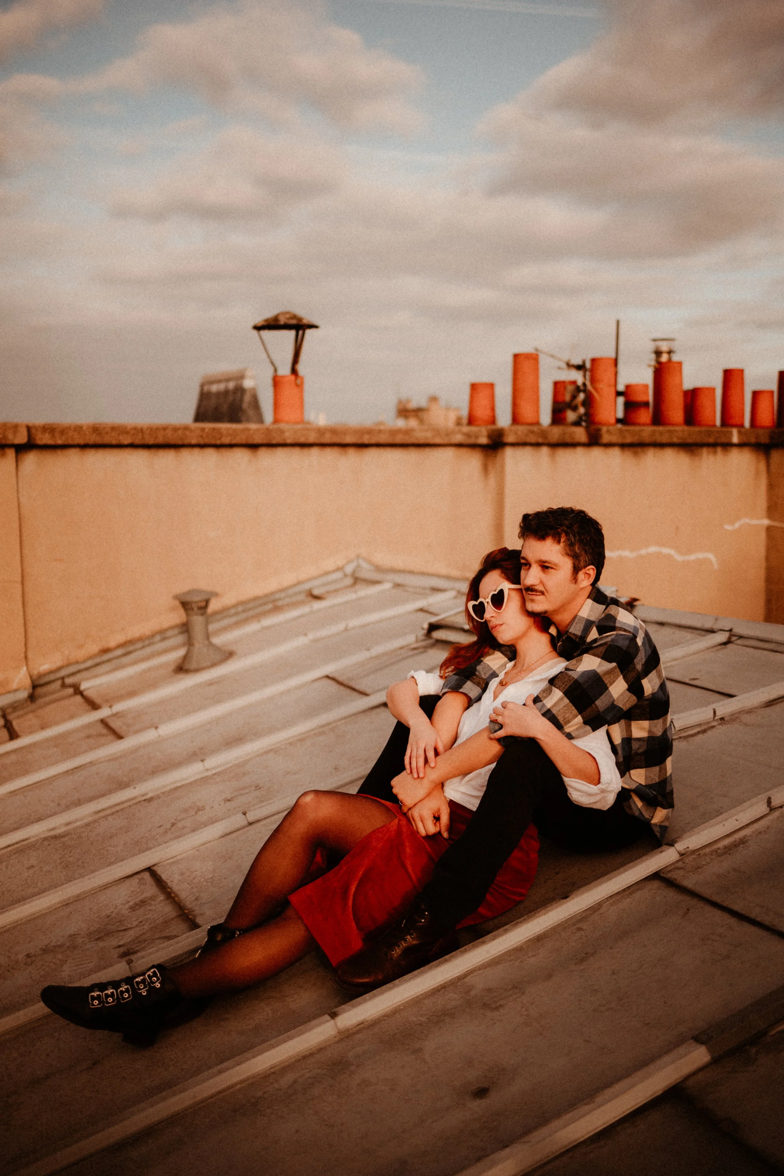 Un couple assis sur un toit à l'heure du coucher du soleil, dans une ambiance romantique, avec un ciel partiellement nuageux en arrière-plan.