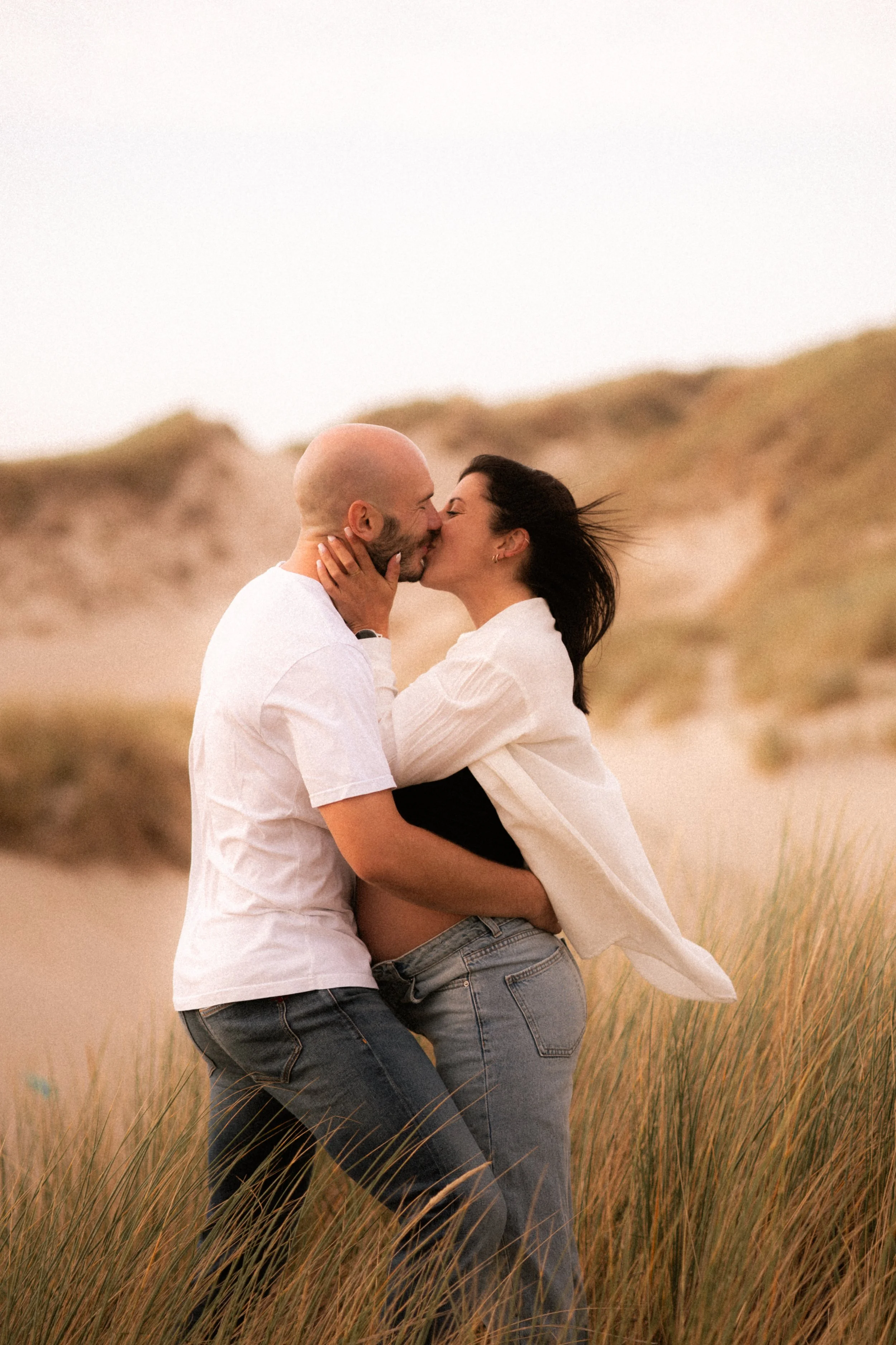 Un couple s'embrasse dans un paysage de dunes et d'herbes hautes lors d'une journée ensoleillée.