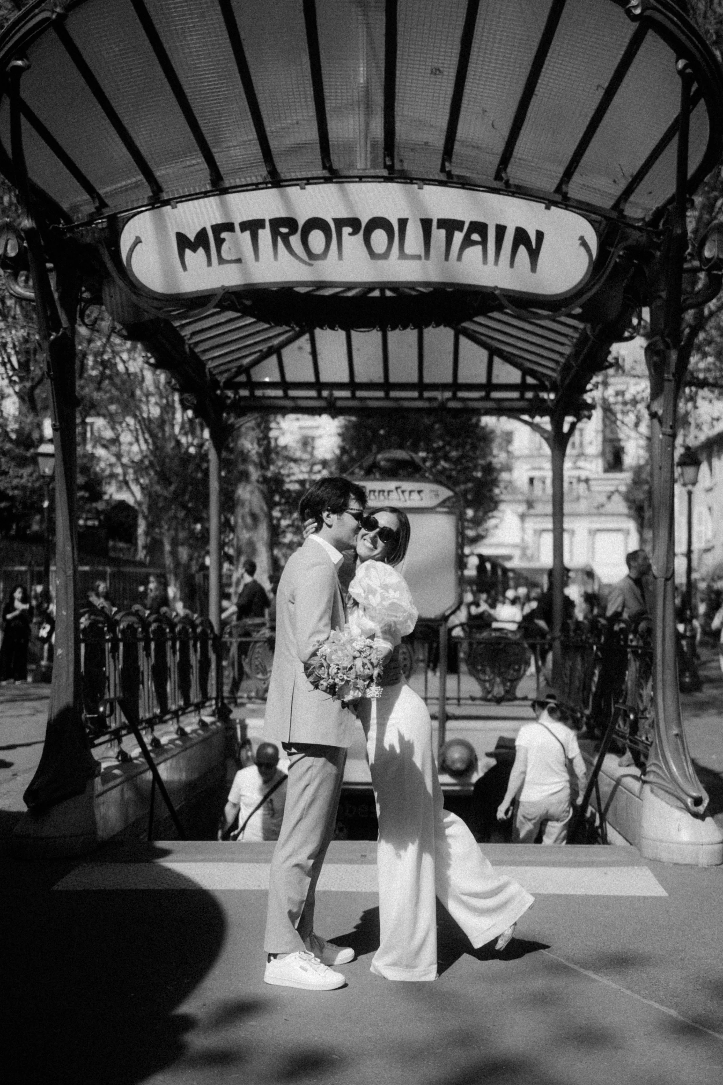 Un couple en mariage, souriant et se tenant par la main, sous une entrée de métro parisien nommé 'Metropolitain'.