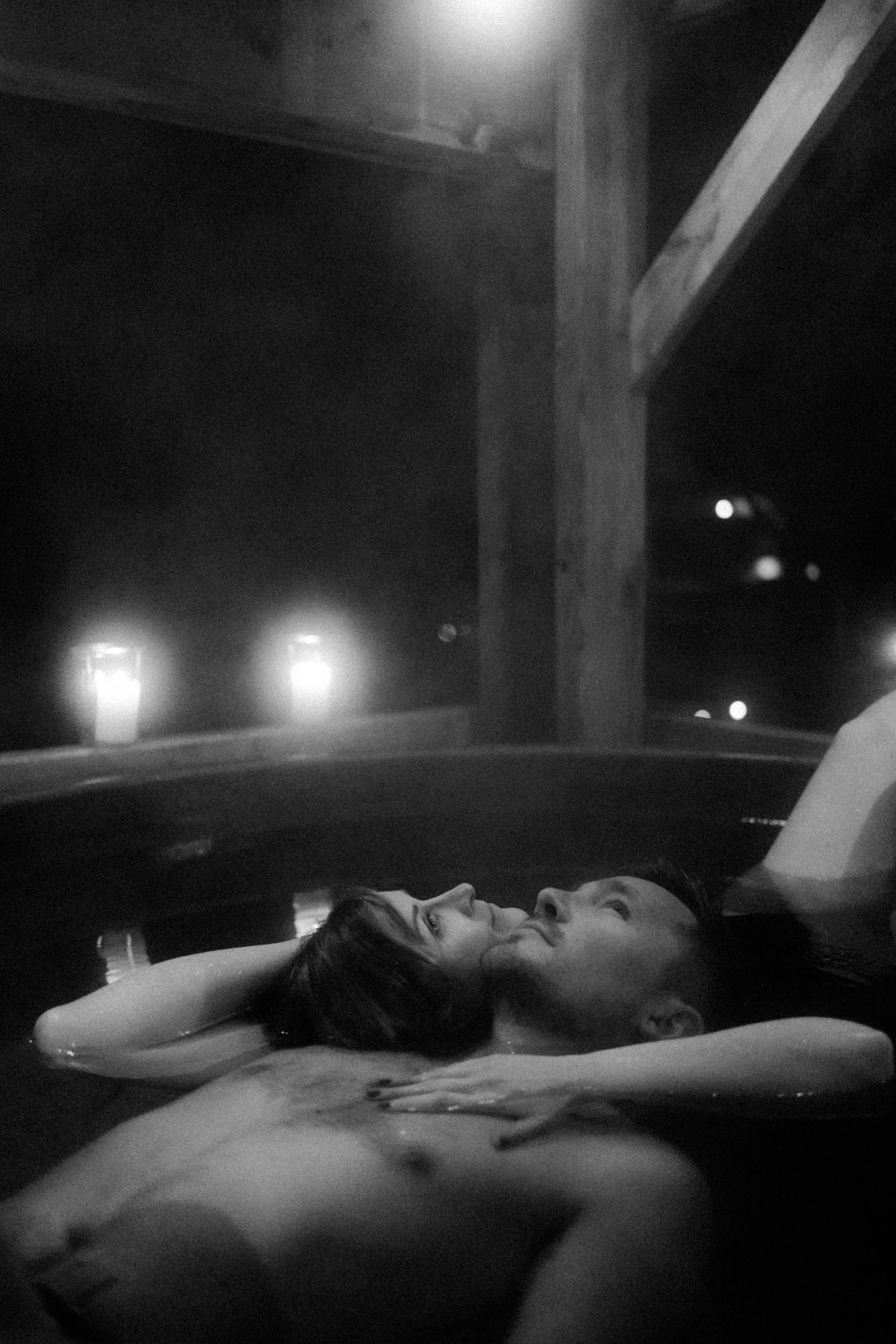 Un couple nue allongé dans une baignoire, regardant vers le plafond, avec une ambiance sombre et mystérieuse avec des lumières floues en arrière-plan.