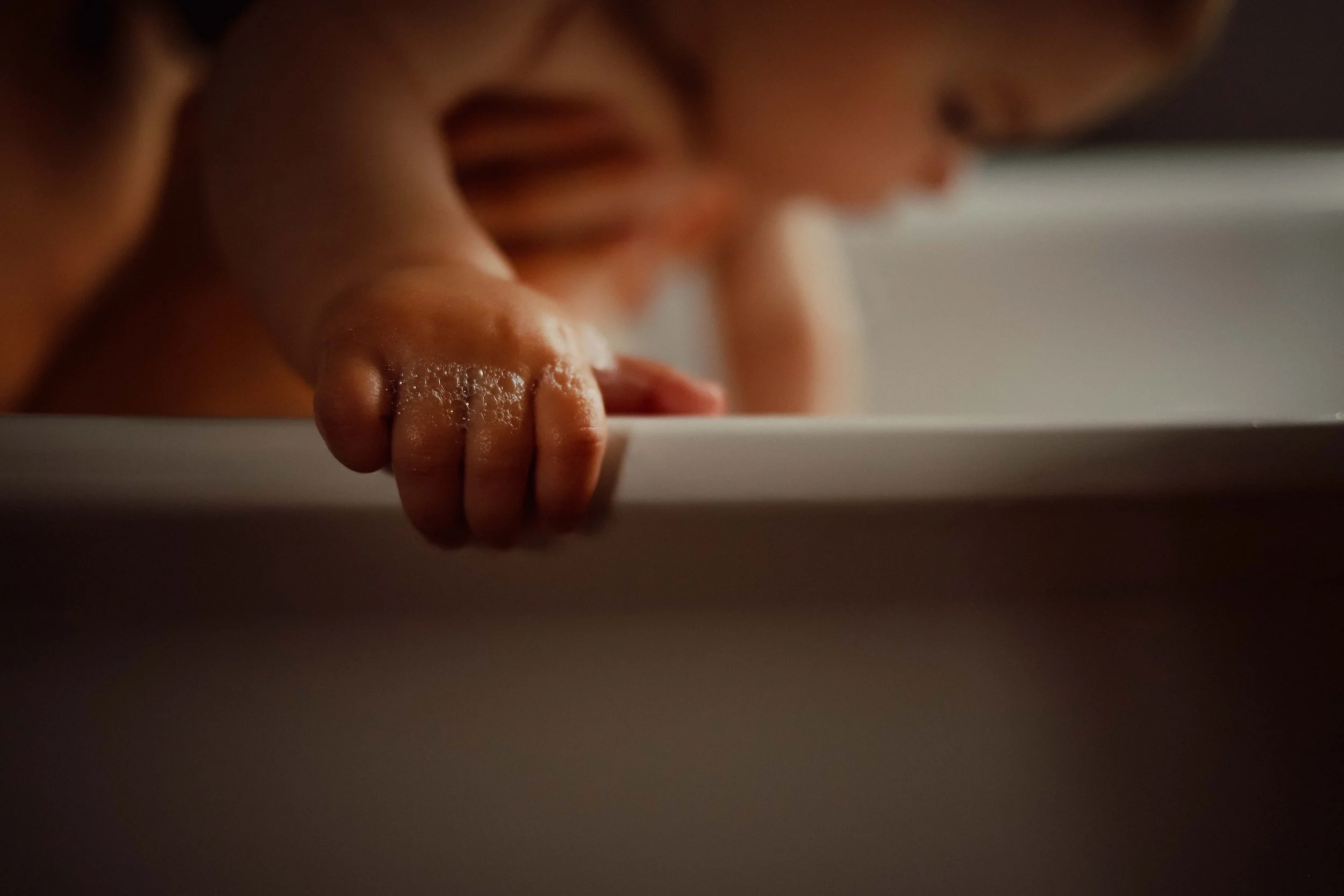 Un bébé qui tient la bordure d'une baignoire, en train de jouer ou de se pencher, avec de l'eau ou du savon sur ses doigts, la scène est floue et sombre.