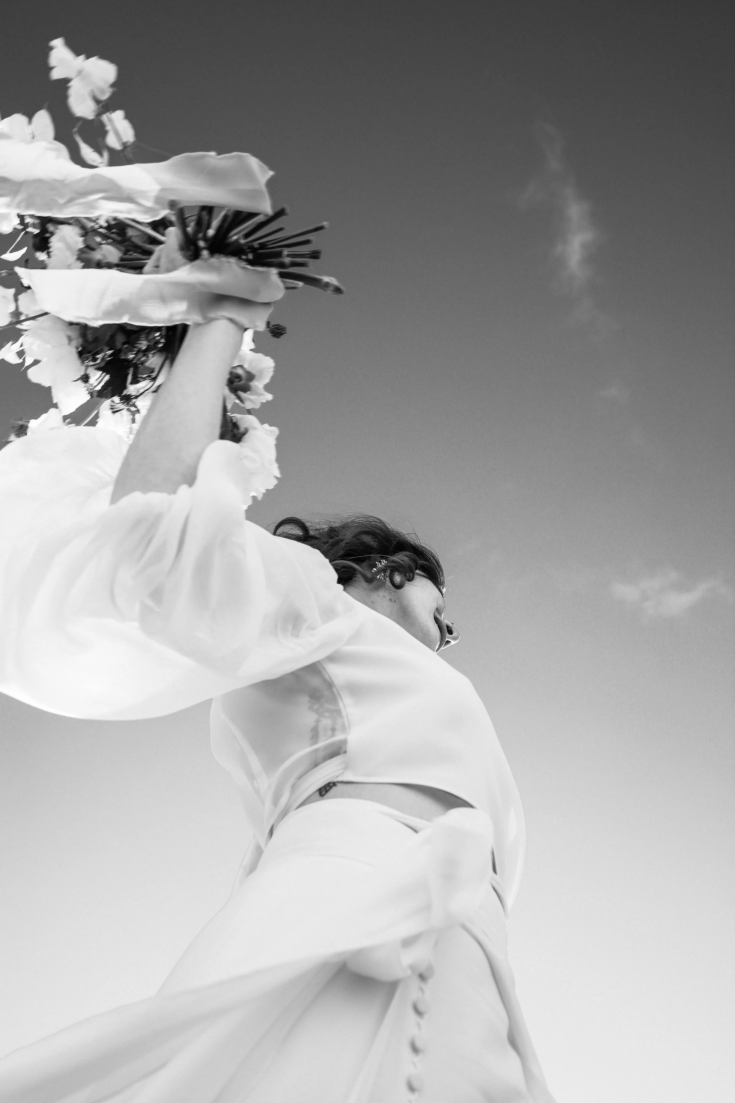 Femme en robe blanche levant un bouquet de fleurs vers le ciel dans un ciel dégagé