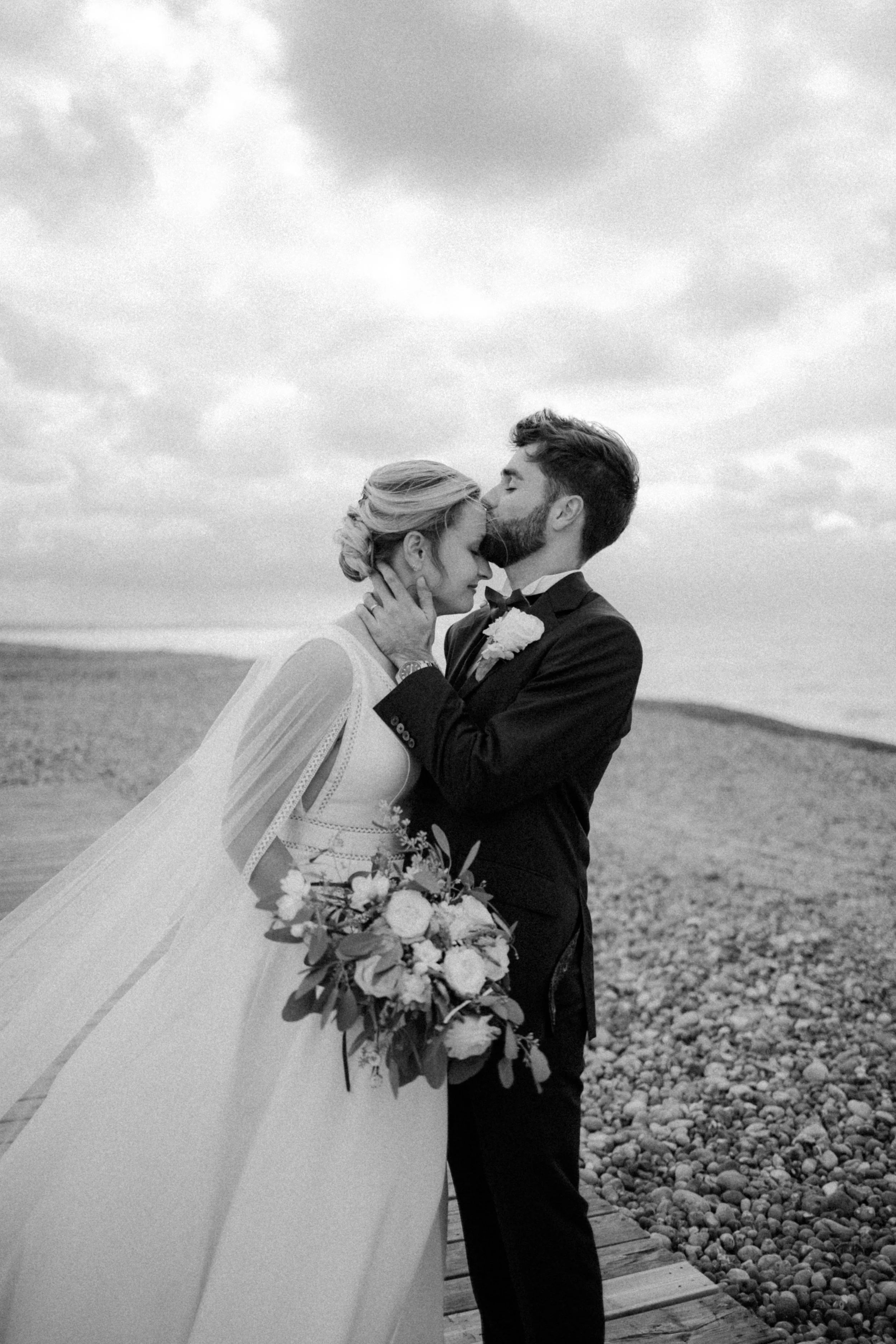 Un couple marié en tenue de mariage, en noir et blanc, se tient sur une jetée en bord de mer, avec des nuages dans le ciel, en train de s'embrasser tendrement.