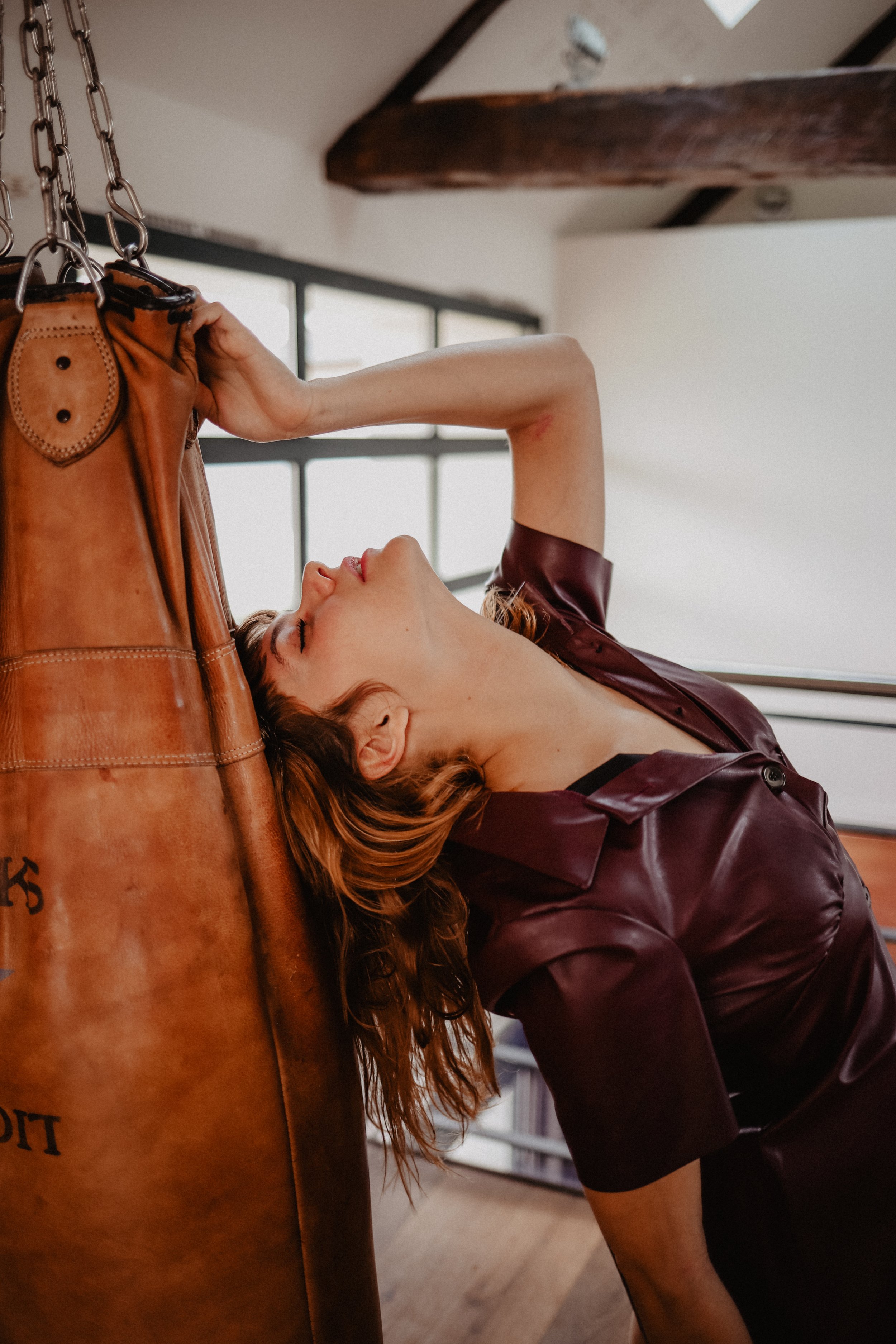 Femme aux cheveux roux en blouse en cuir marron, posant avec la tête inclinée en arrière contre un sac de boxe en cuir