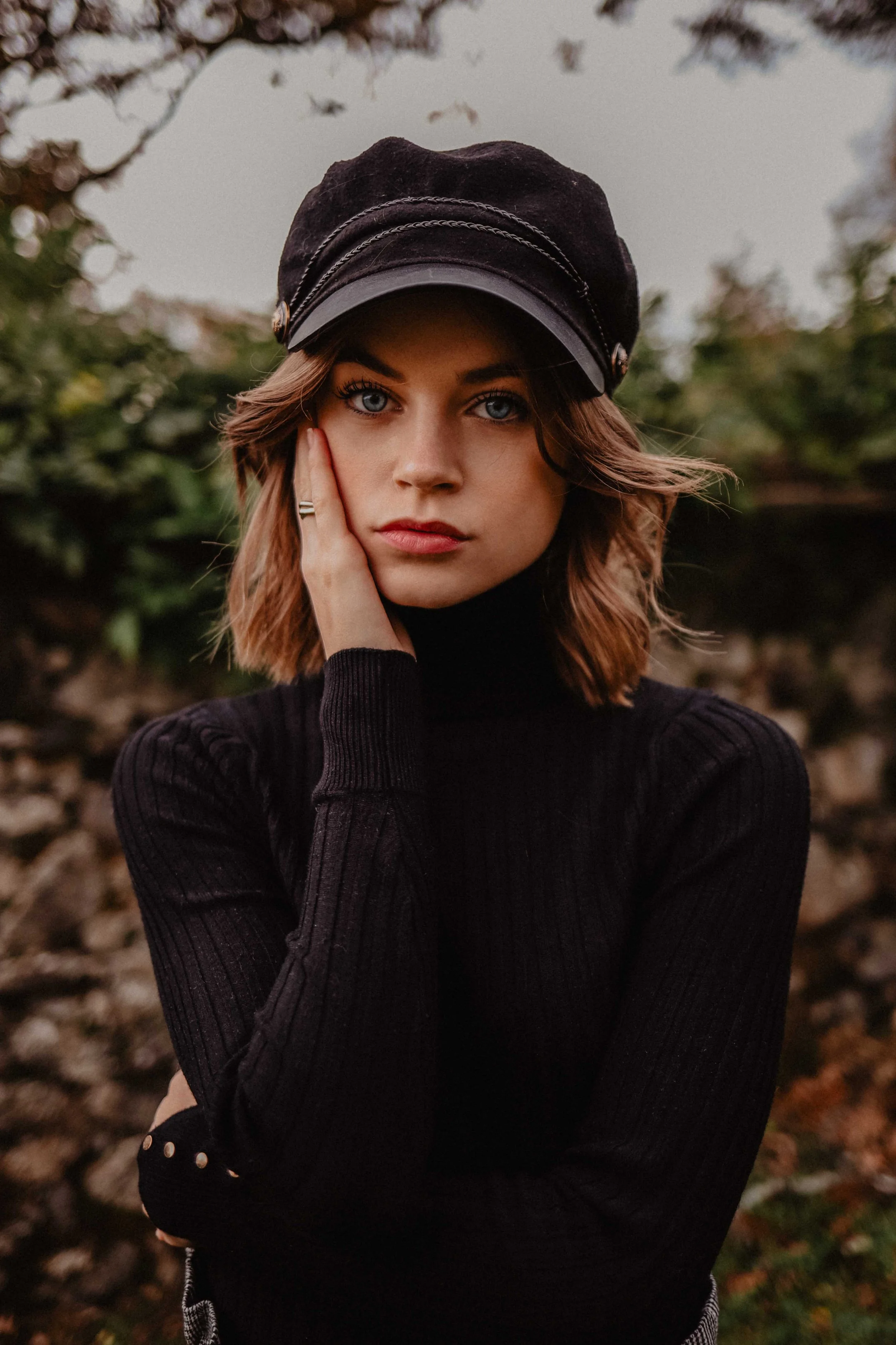 Une jeune femme aux cheveux bruns, portant un bonnet noir, une robe ou pull noir à col roulé, pose en regardant l'objectif avec un fond flou d'arbres et de végétation.