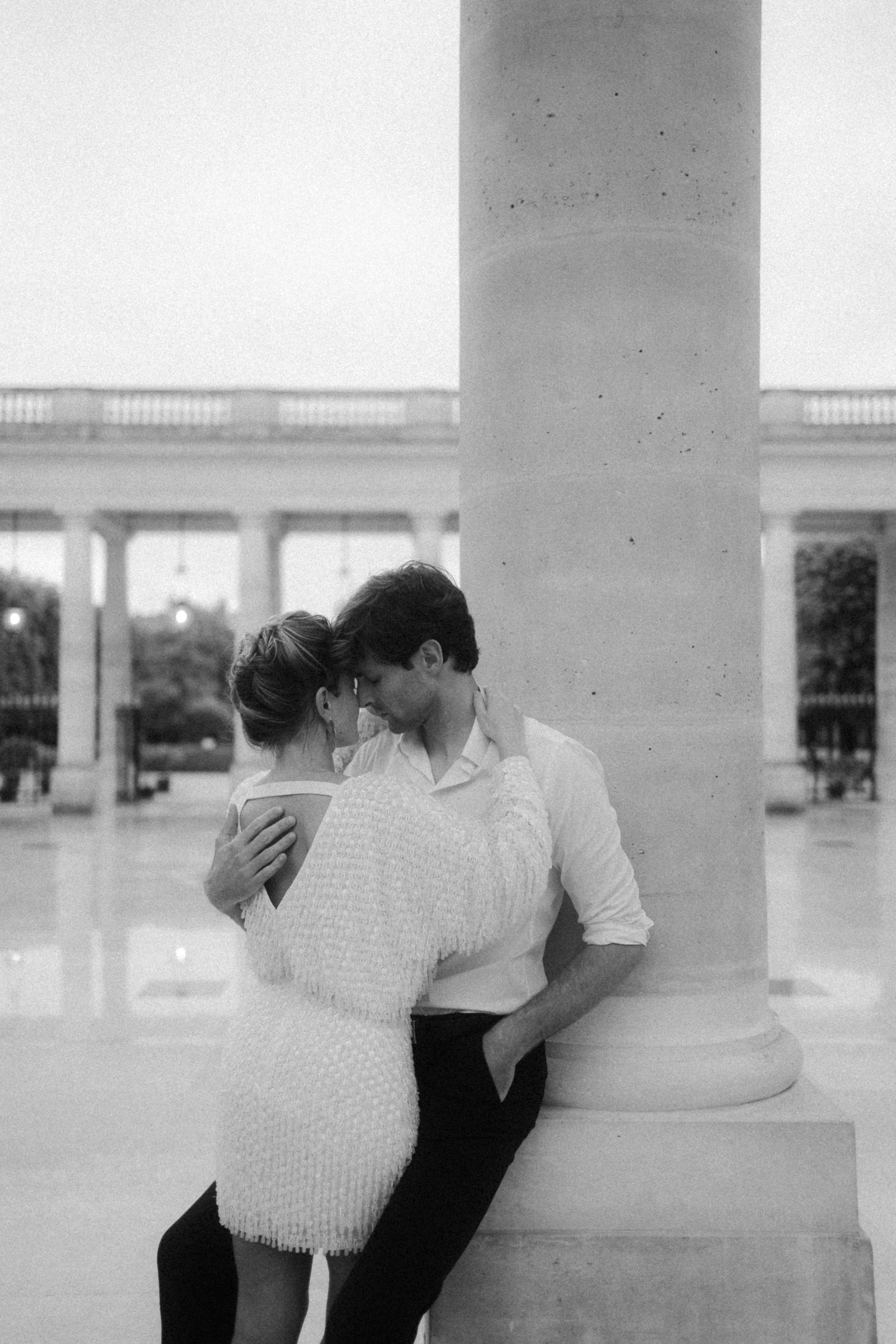 Un couple s'enlace près d'une colonne dans un lieu public extérieur, en noir et blanc.
