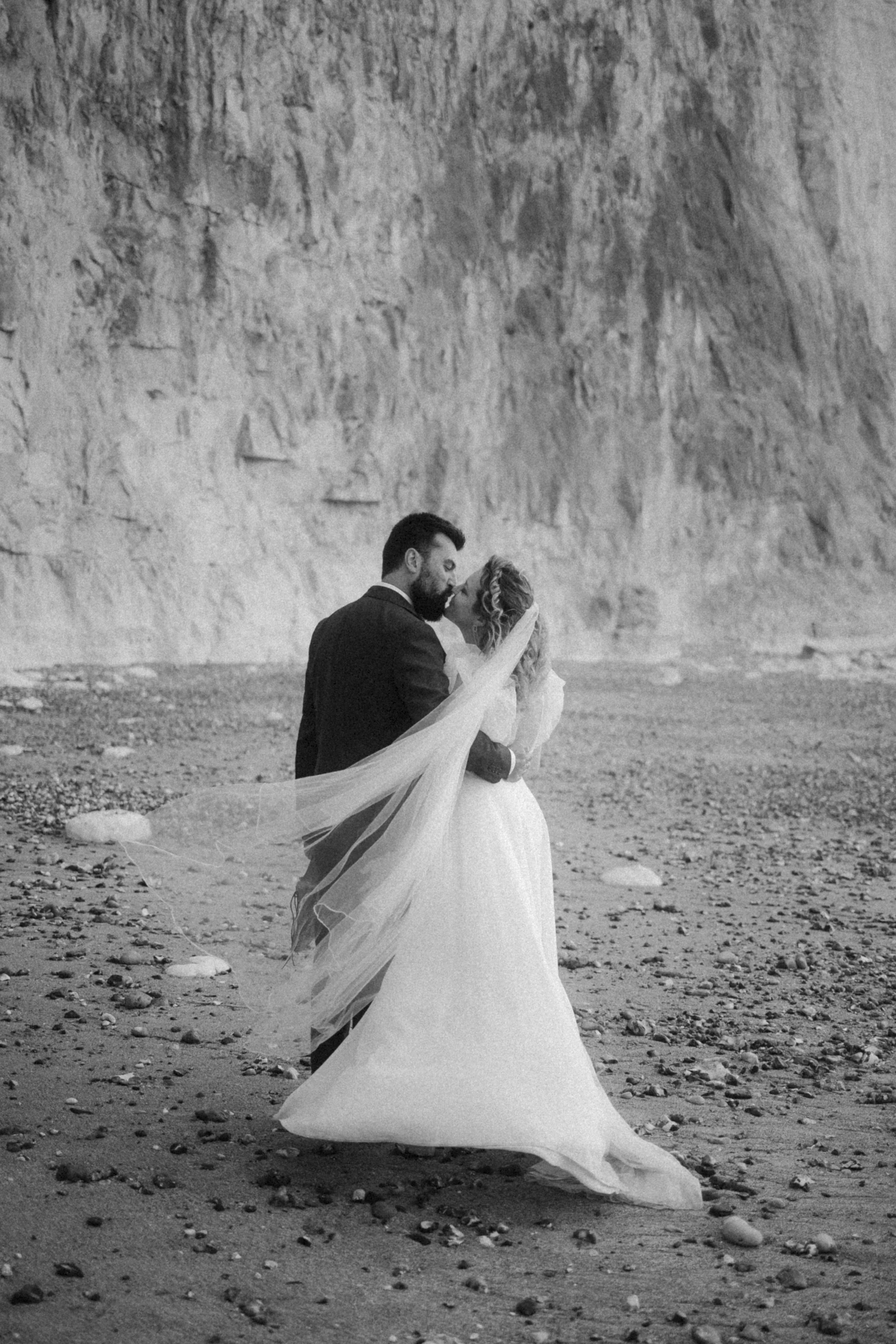 Un couple de mariés s'embrassant sur une plage avec des falaises en arrière-plan, en noir et blanc.