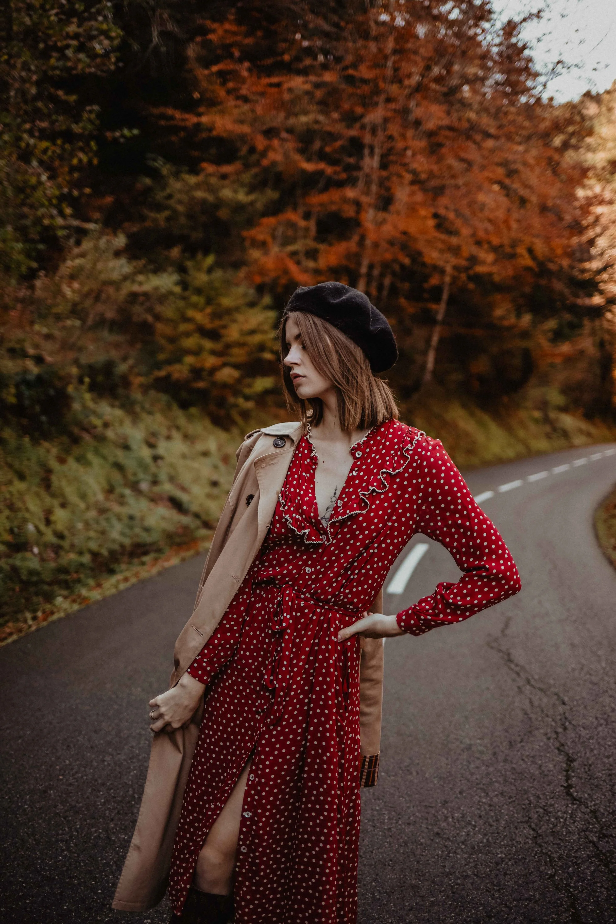Une jeune femme portant une robe rouge à pois blancs, un trench beige à ses épaules, et un béret noir, se tient sur une route serpentée au milieu d'une forêt automnale aux arbres aux feuilles orange et marron.