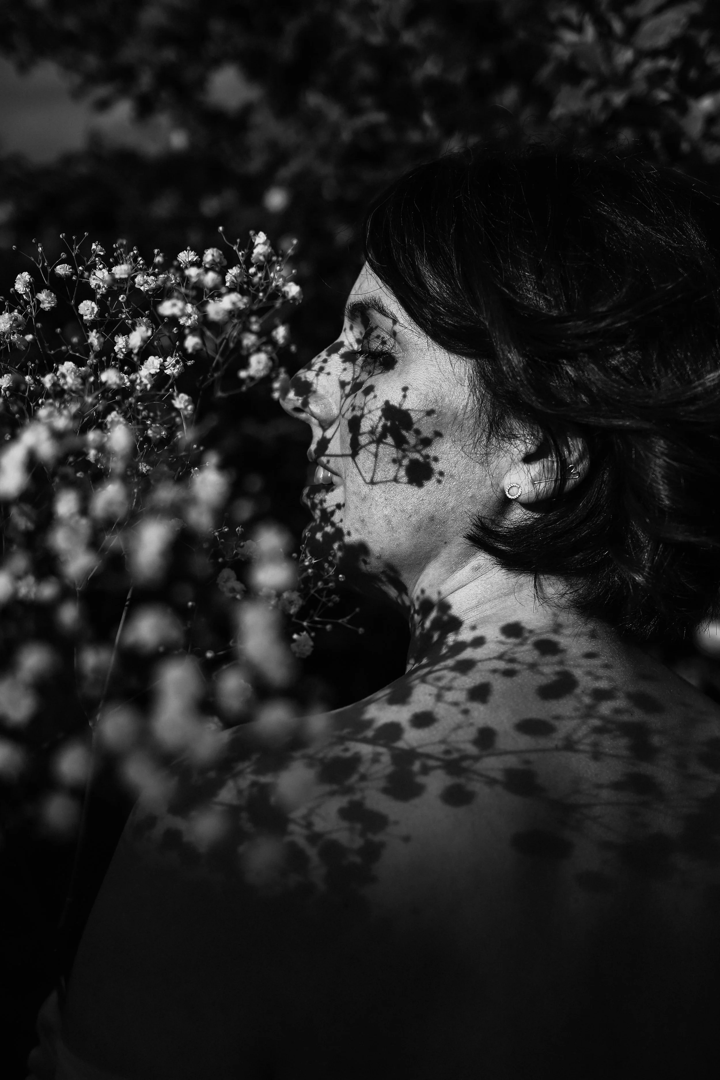 Une femme avec des cheveux courts regarde une branche de petites fleurs. L'ombre des fleurs projette un motif sur son visage, son épaule et son cou, dans une photographie en noir et blanc.