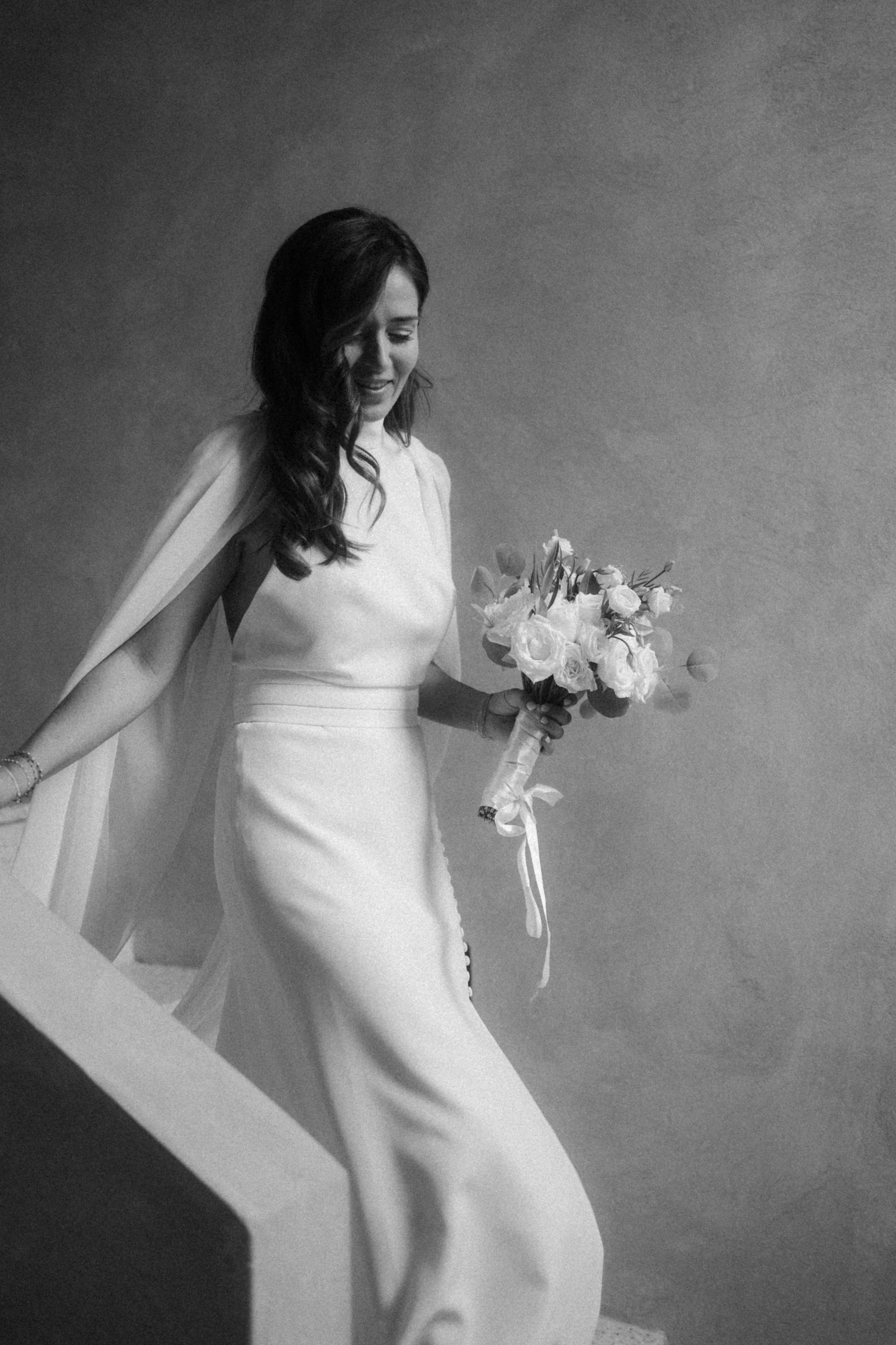 Une femme en robe blanche de mariage, tenant un bouquet de fleurs, souriante et marchant dans une ambiance intime.