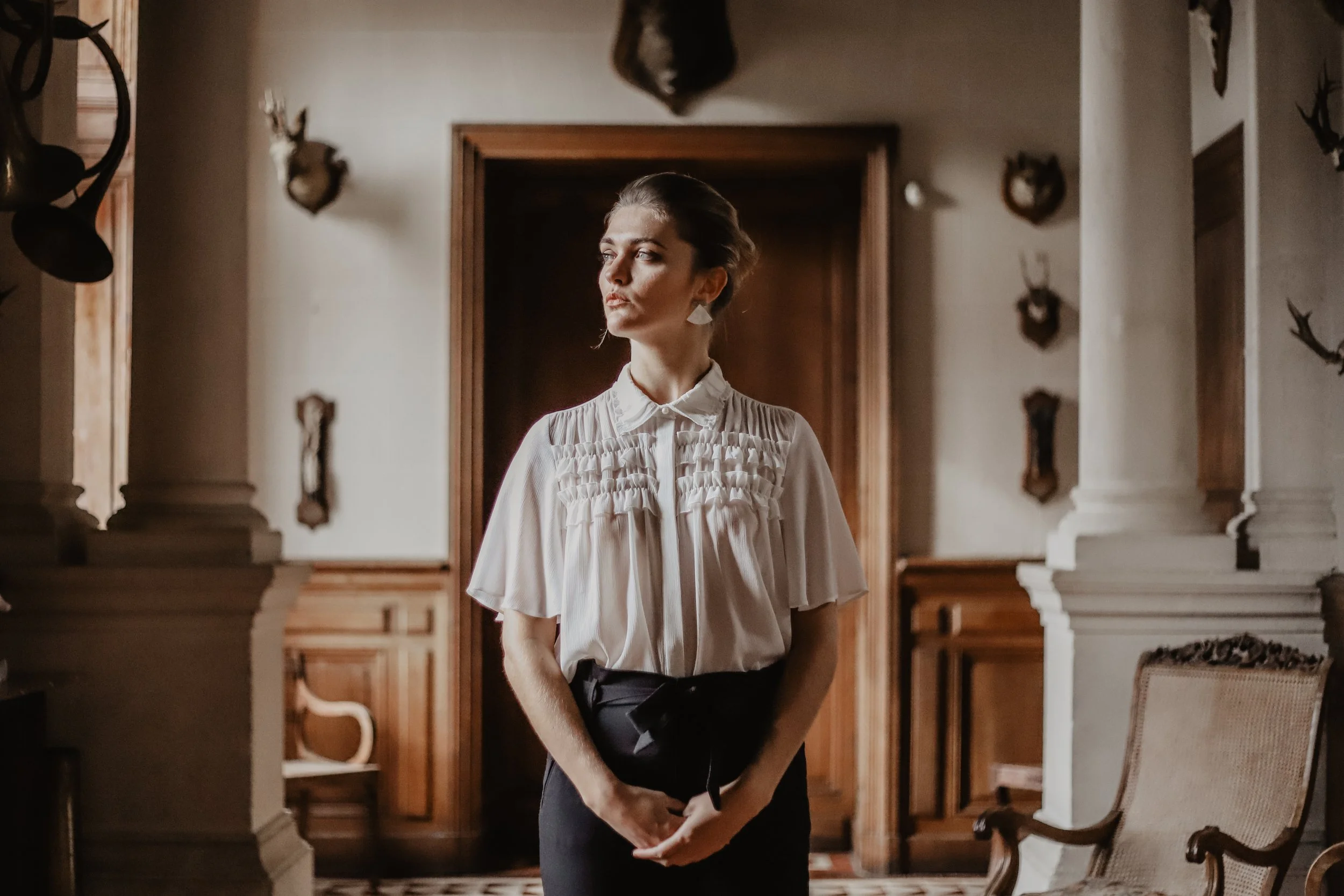Une jeune femme debout dans une pièce décorée avec des trophées de chasse au mur, portant une chemise blanche et une jupe noire.