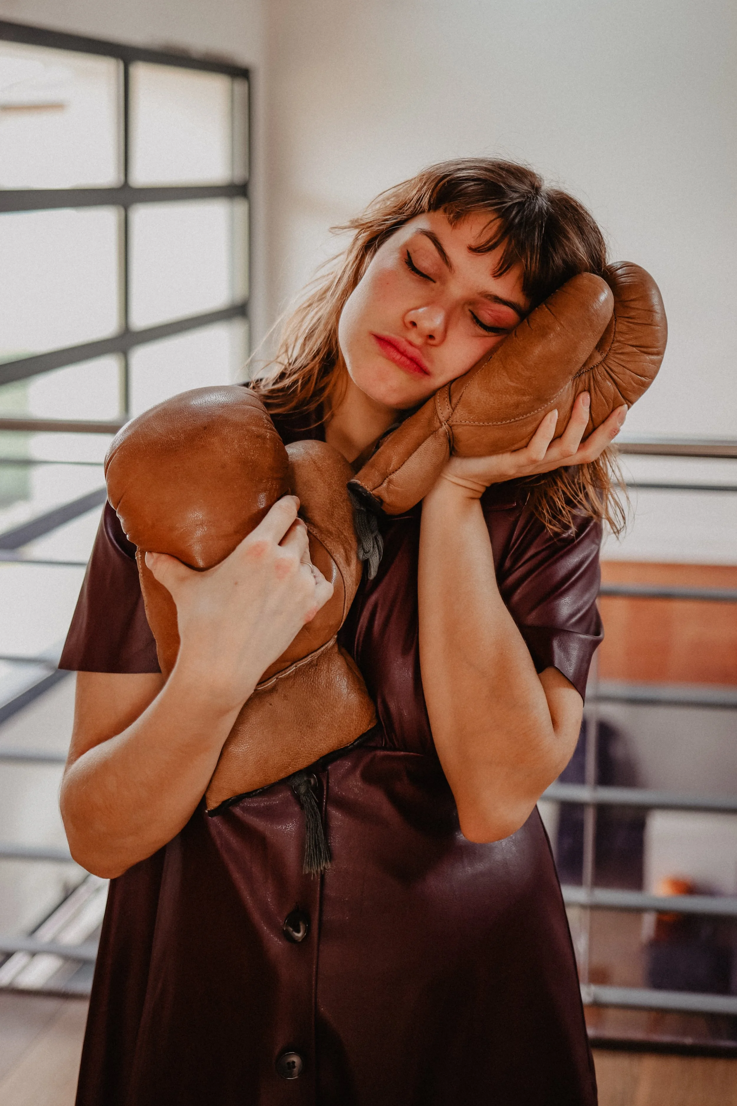 Une femme avec des gants de boxe marron, portant un robe en cuir bordeaux, ferme les yeux et semble épuisée ou rêveuse dans un espace intérieur moderne.