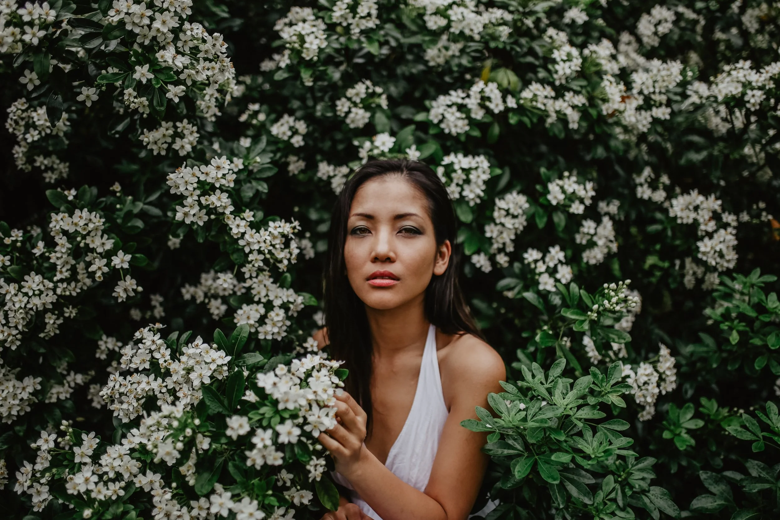 Femme avec des cheveux foncés portant une robe blanche, entourée de buissons de fleurs blanches et de feuilles vertes.