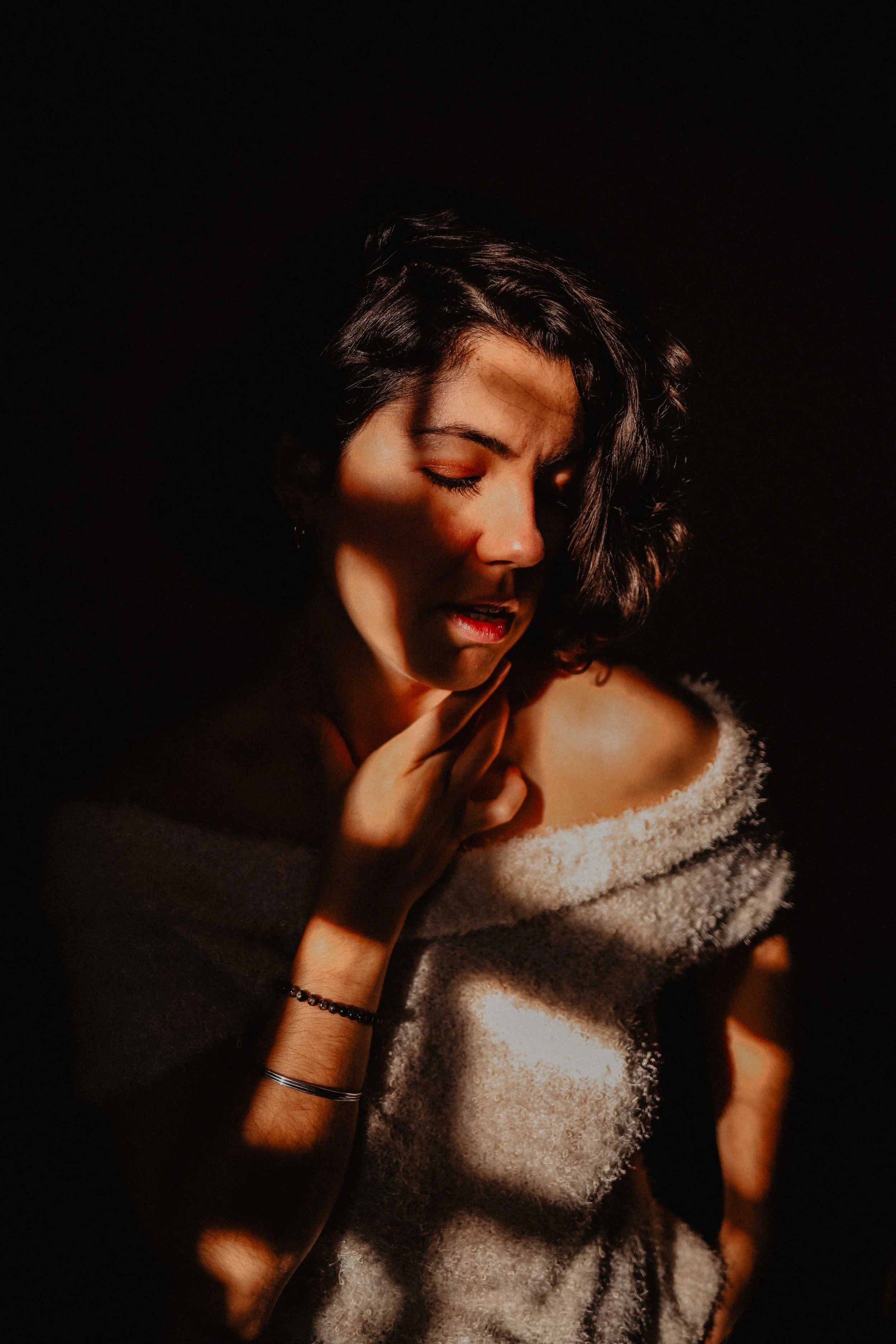 Une femme avec des cheveux bouclés courts, portant un pull en laine, se tient dans une lumière tamisée, avec une expression pensive et contemplative.