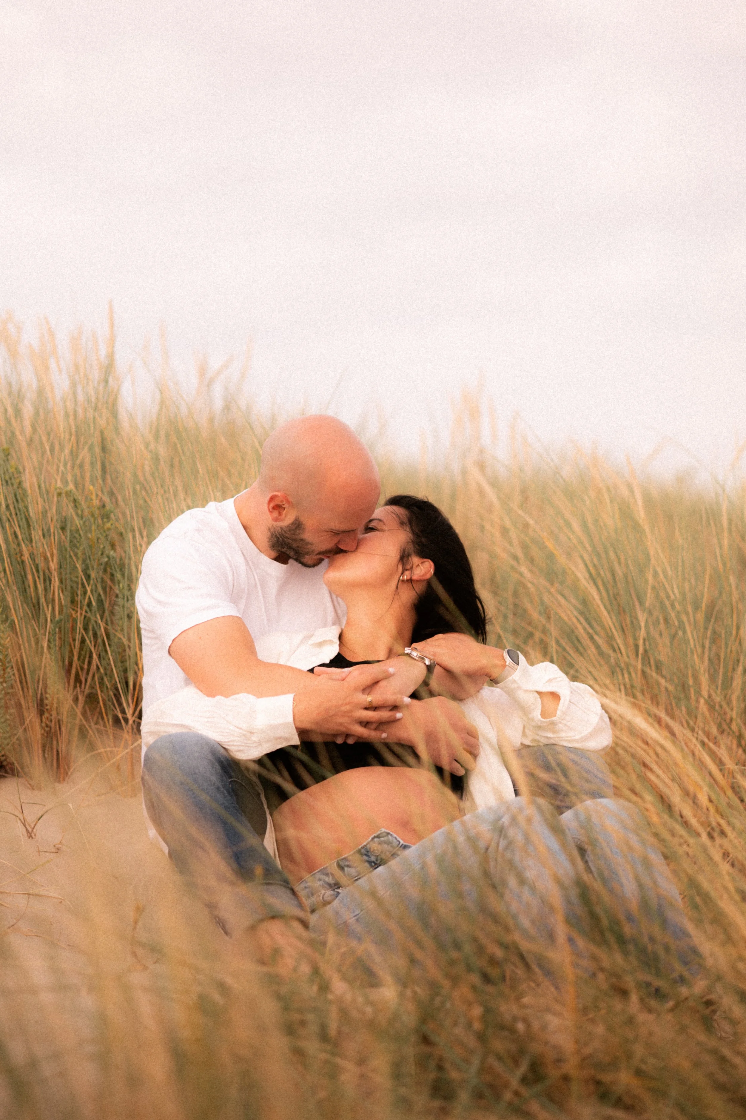 Un couple en embrasse sur une plage avec des herbes hautes, partageant un baiser romantique.