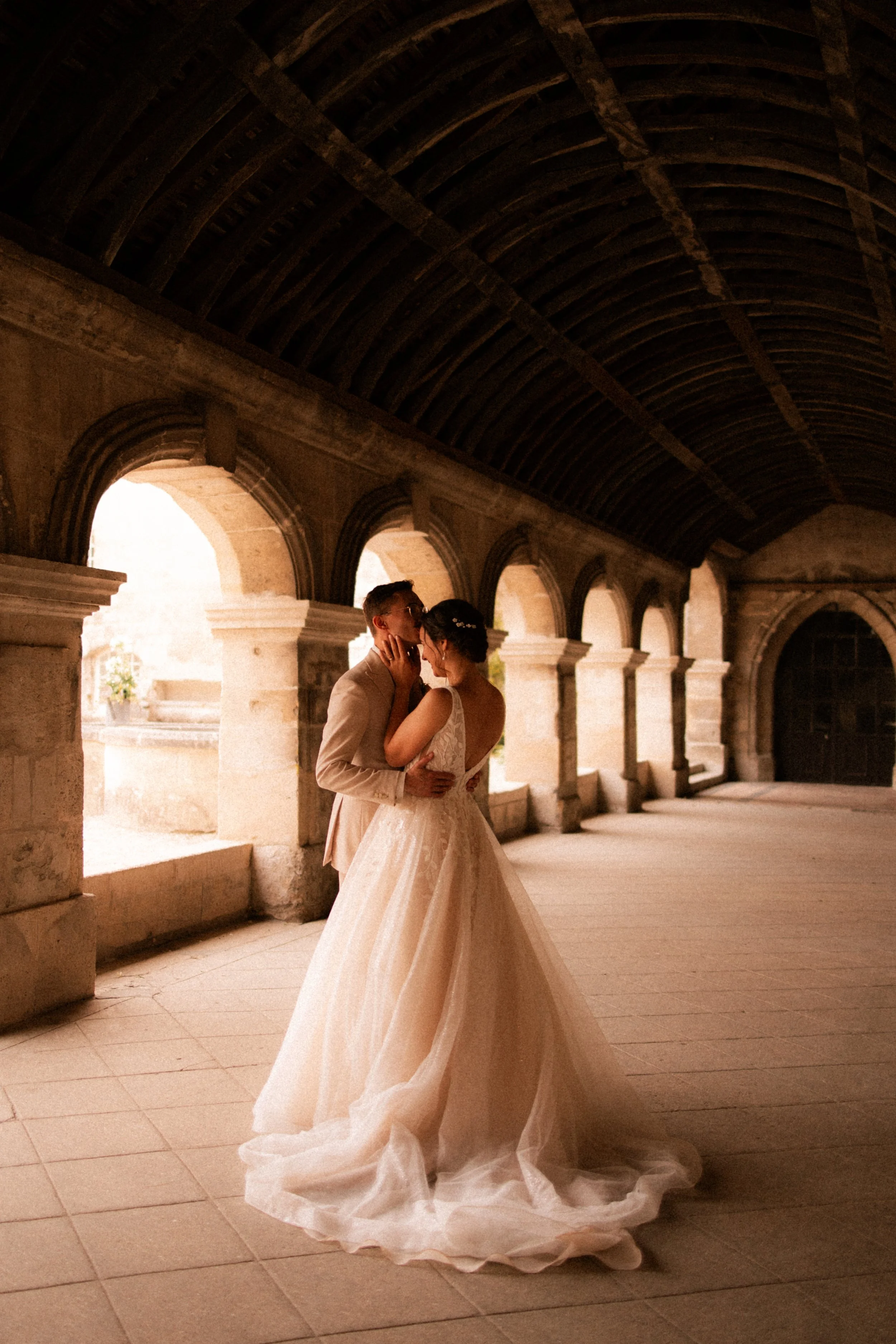 Un couple de mariés s'embrassant dans un couloir voûté en pierre, lumière douce venant de fenêtres ouvertes.