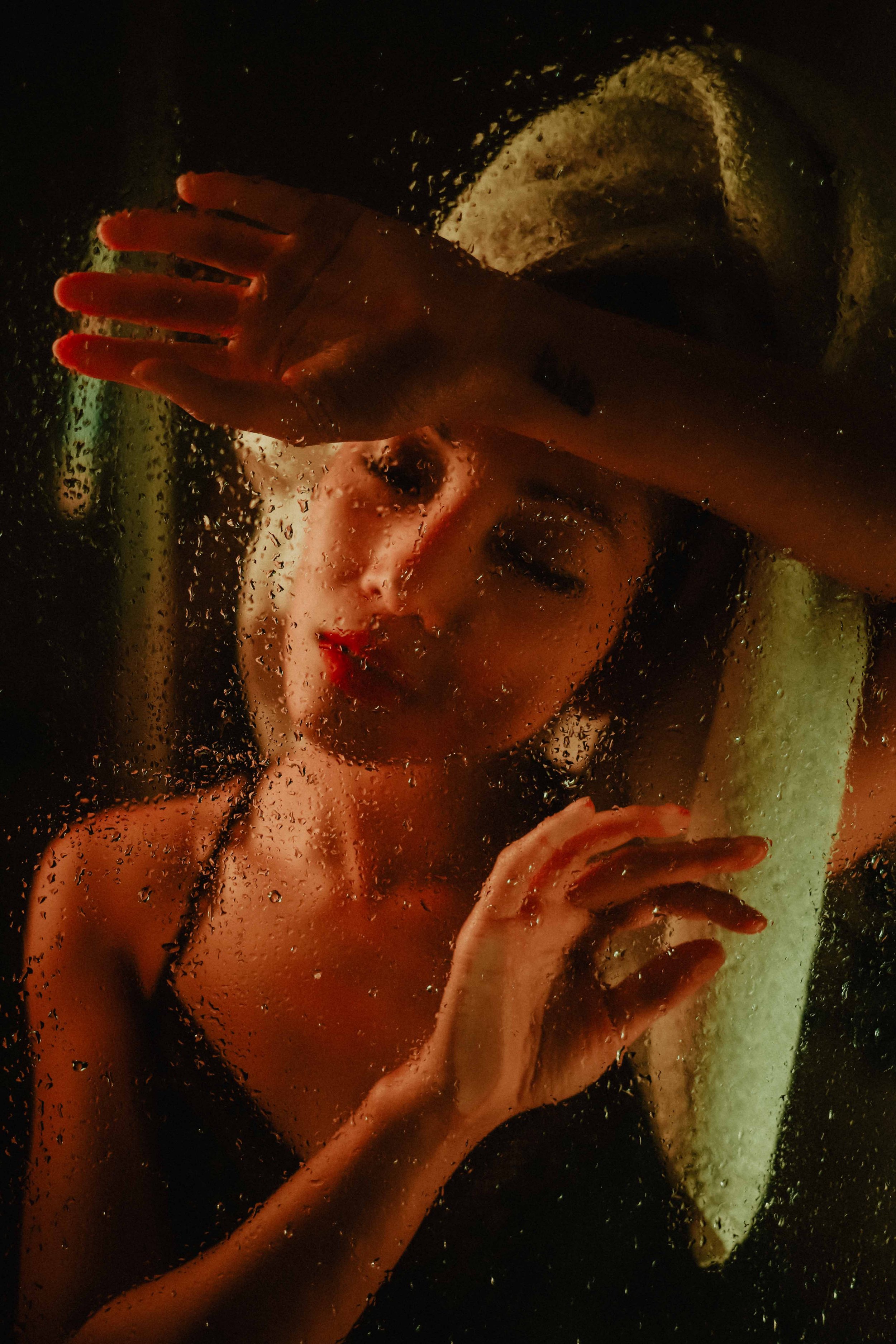 Femme derrière une vitre de douche mouillée, enroulée dans une serviette, yeux fermés, dans une ambiance chaude et intime.