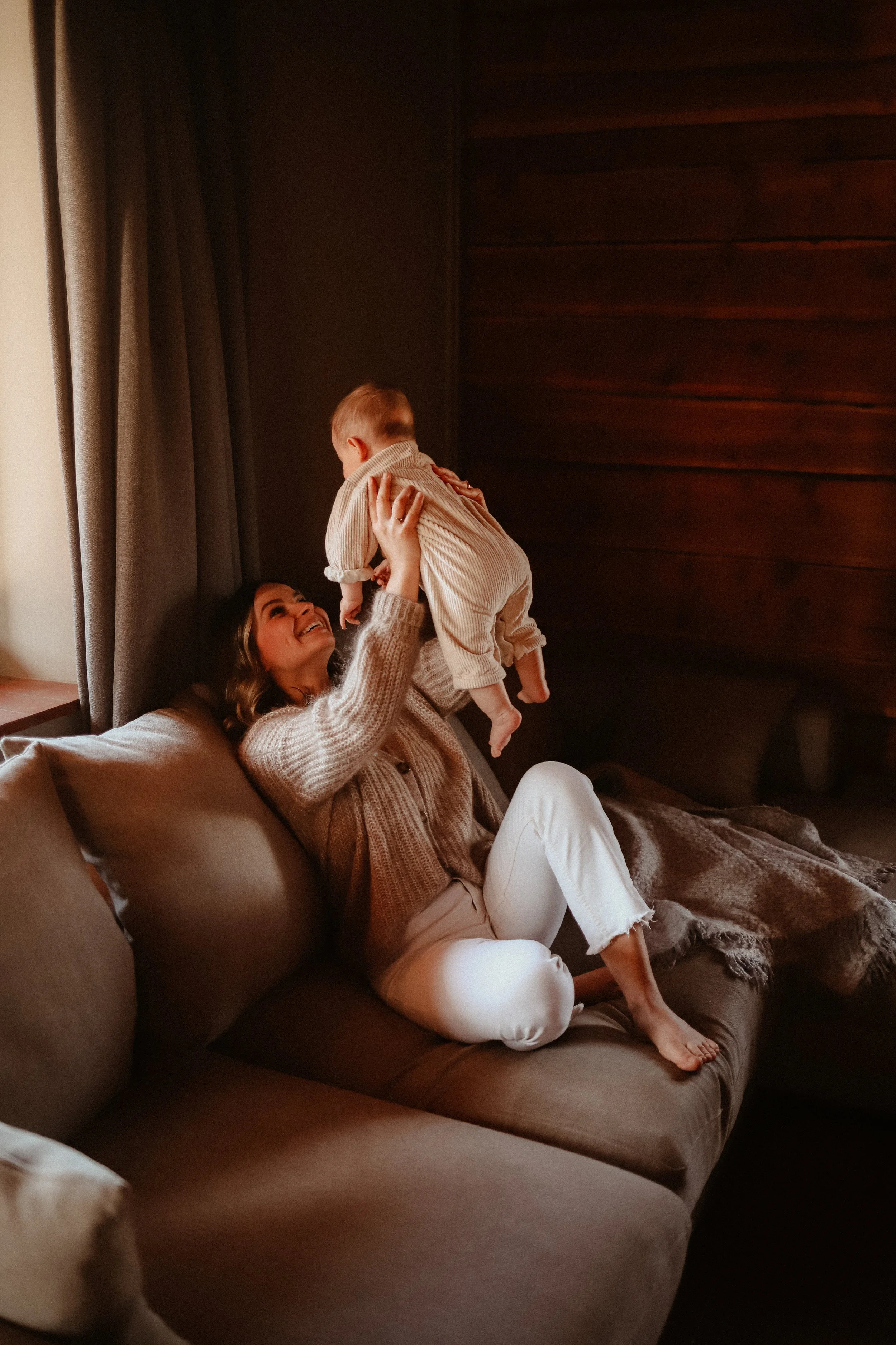 Une femme et un bébé jouent sur un canapé dans un salon chaleureux avec des rideaux gris et des murs en bois.