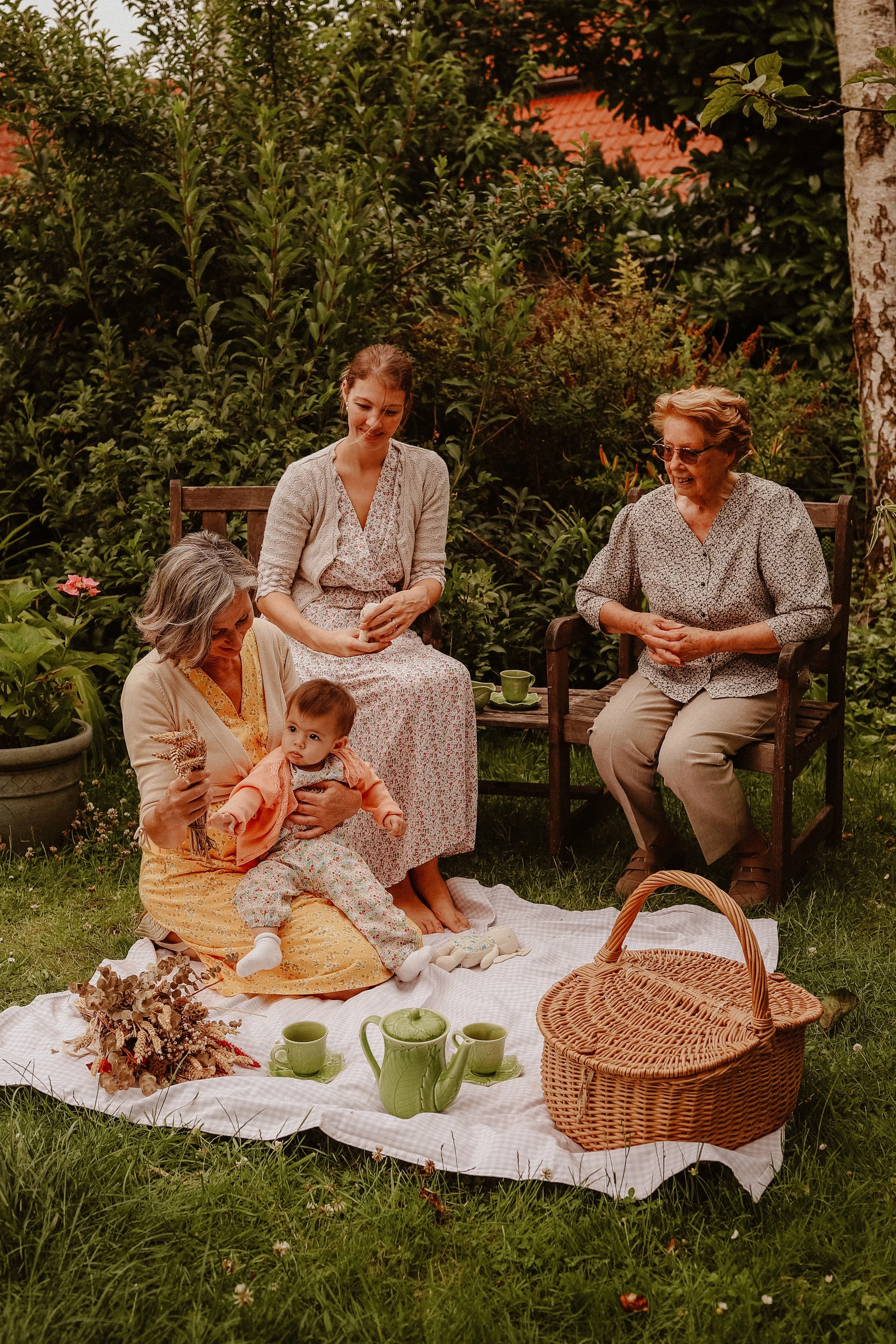 Une famille fait un pique-nique dans un jardin verdoyant, avec plusieurs femmes assises et un bébé sur une nappe, entourée de plantes et de nature.