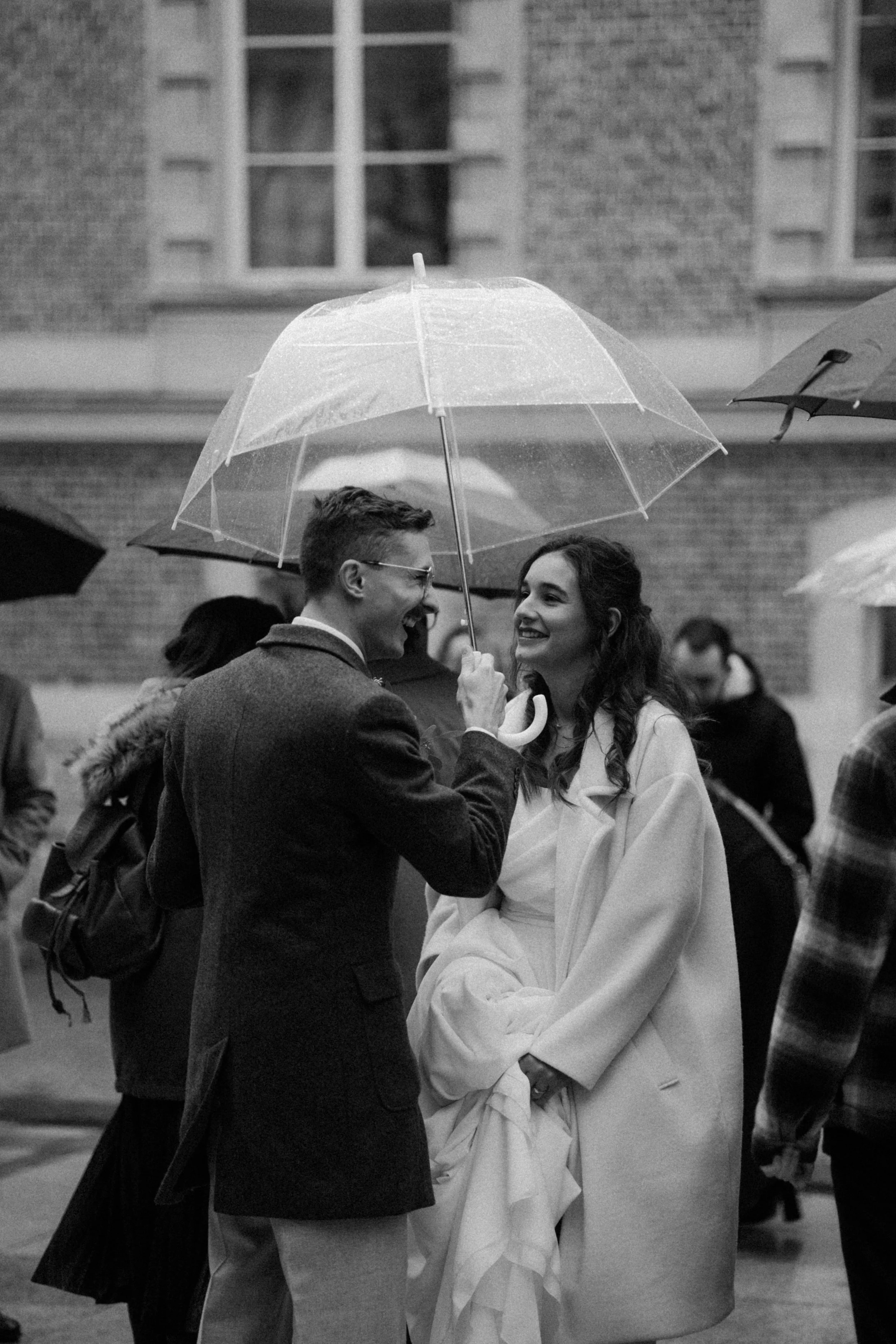 Deux jeunes adultes heureux partageant un moment sous un parapluie dans une rue bondée, en noir et blanc.