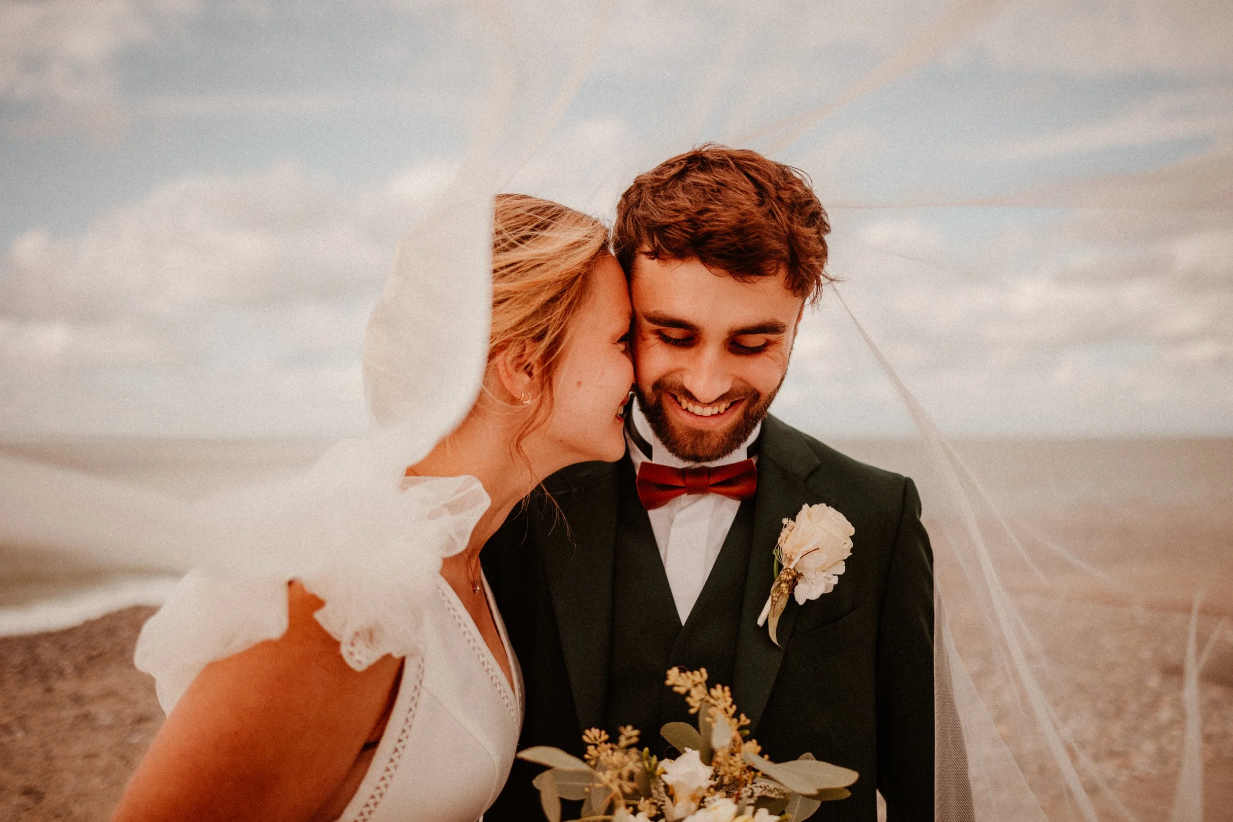 Un couple de mariés souriant sous un voile de mariée lors d'un mariage en plein air, avec un ciel nuageux en arrière-plan.