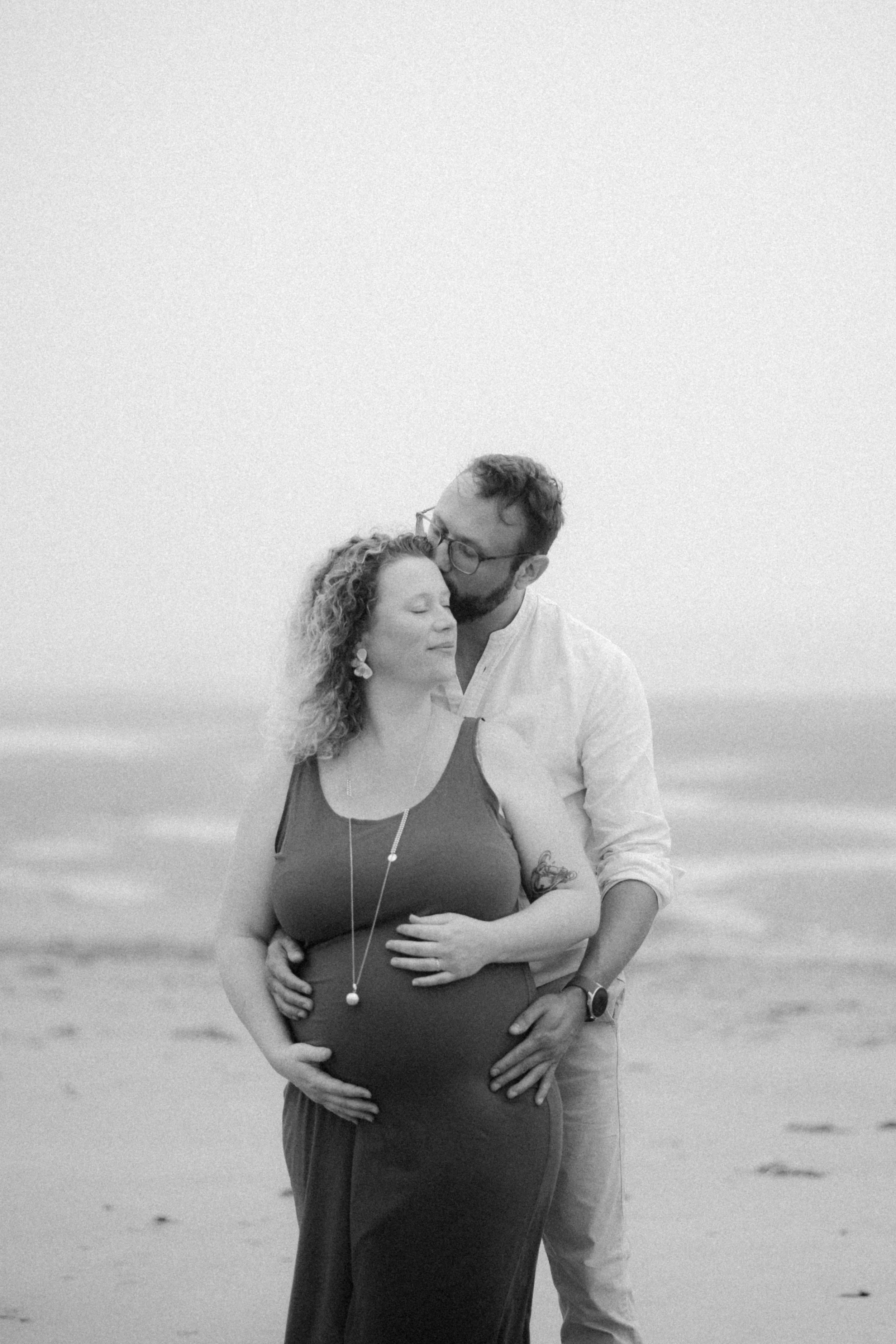 Un couple en détente sur la plage, l'homme embrasse la femme enceinte dans un décor marin.
