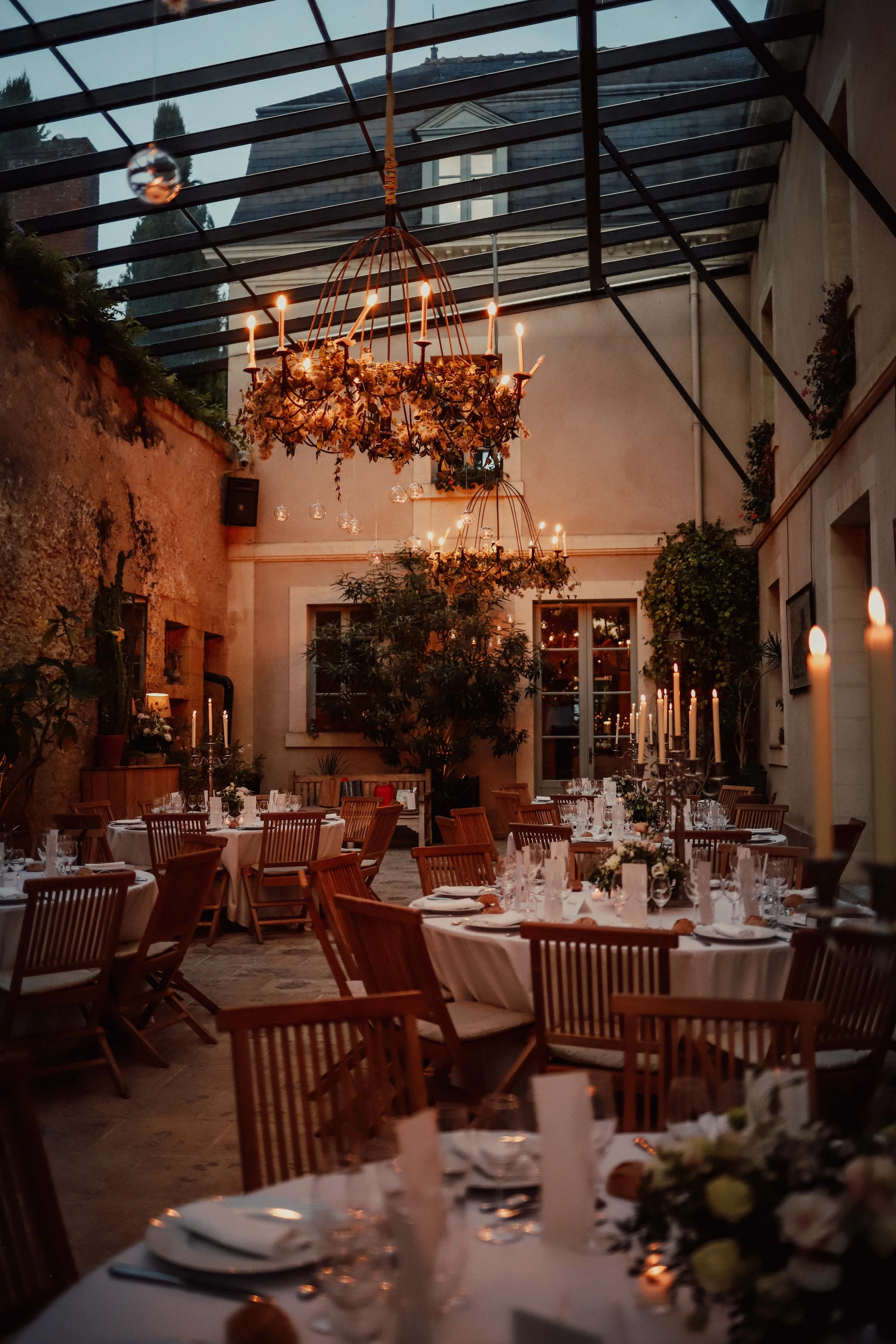 Salle à manger décorée pour un événement, avec des chandelles, des tables rondes avec des nappes blanches, des fleurs, et des chandeliers, sous un plafond en verre avec des lustres de fleurs et des bougies.