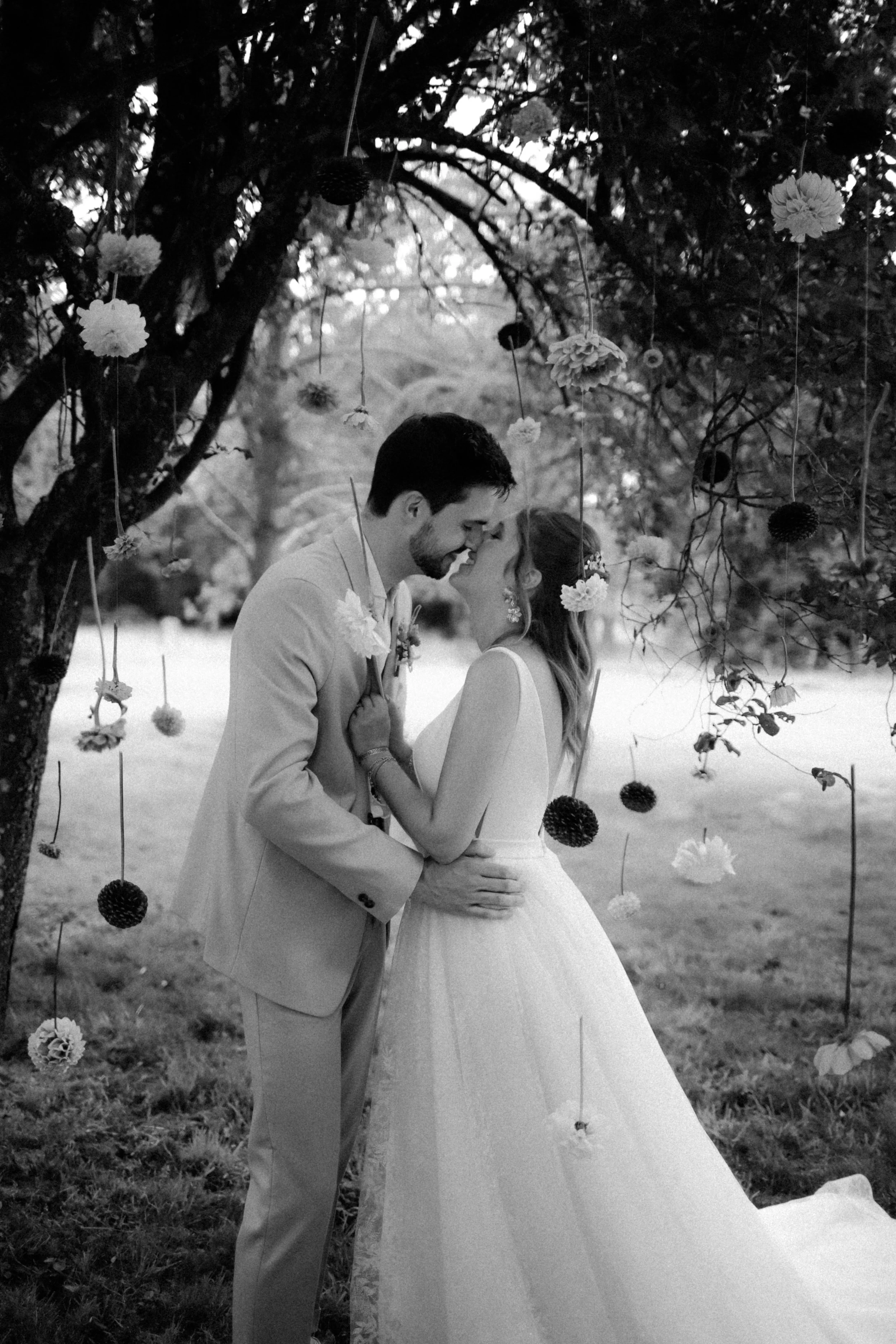 Un couple de mariés embrassant sous un arbre décoré de fleurs et de boules suspendues, en noir et blanc.