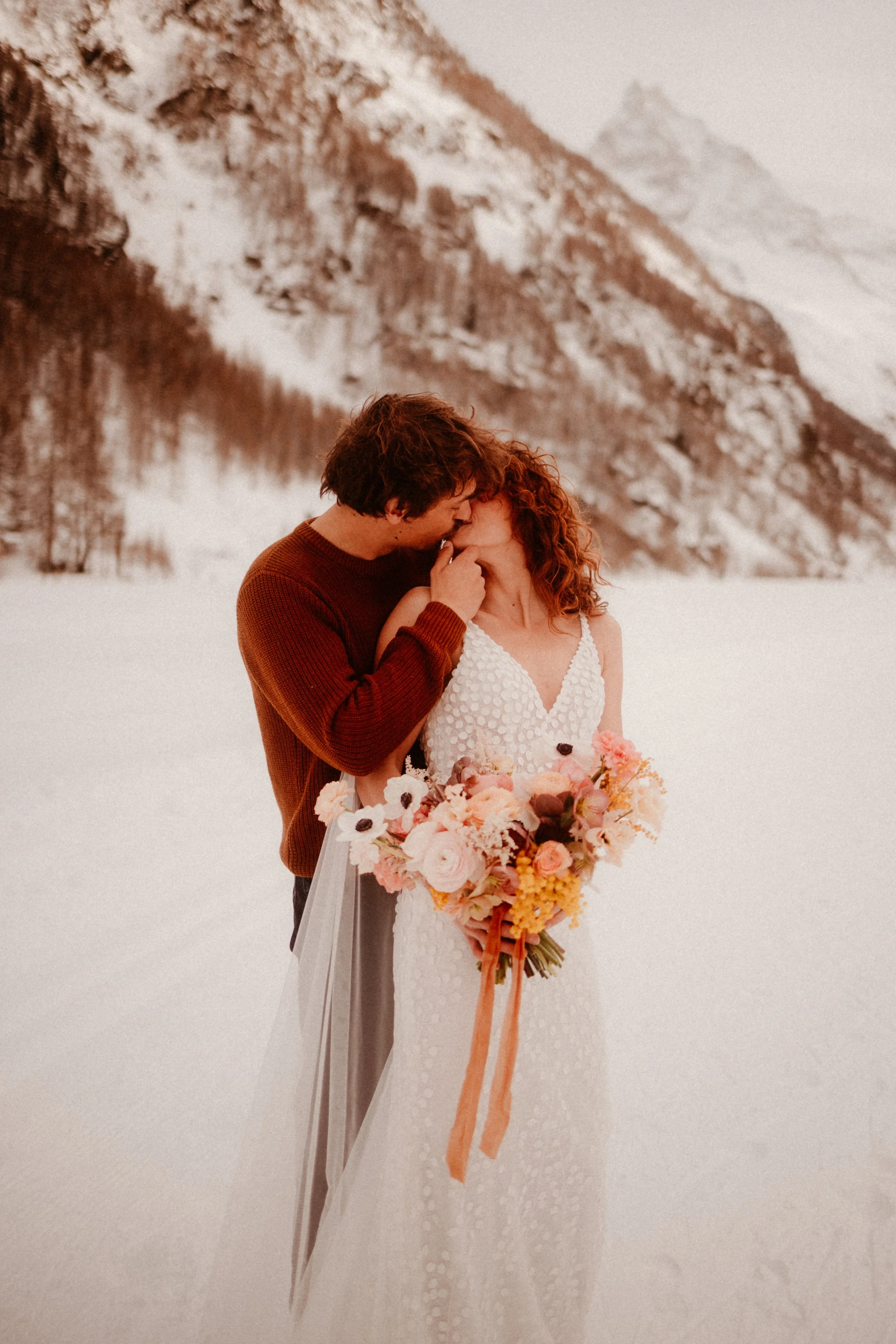 Un couple qui s'embrasse dans la neige, la femme tenant un bouquet de fleurs, en arrière-plan des montagnes enneigées.