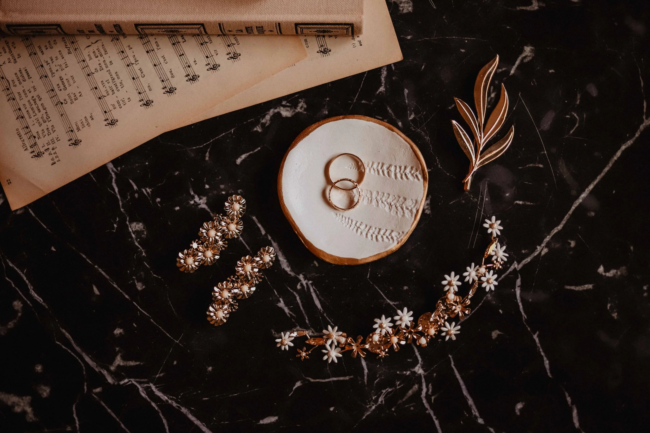 Des bijoux de mariage, comprenant des anneaux, des boucles d'oreilles en fleurs, un bracelet avec des marguerites et une broche en forme de branche, posés sur une surface en marbre noir. En arrière-plan, un livre ouvert avec de la musique.