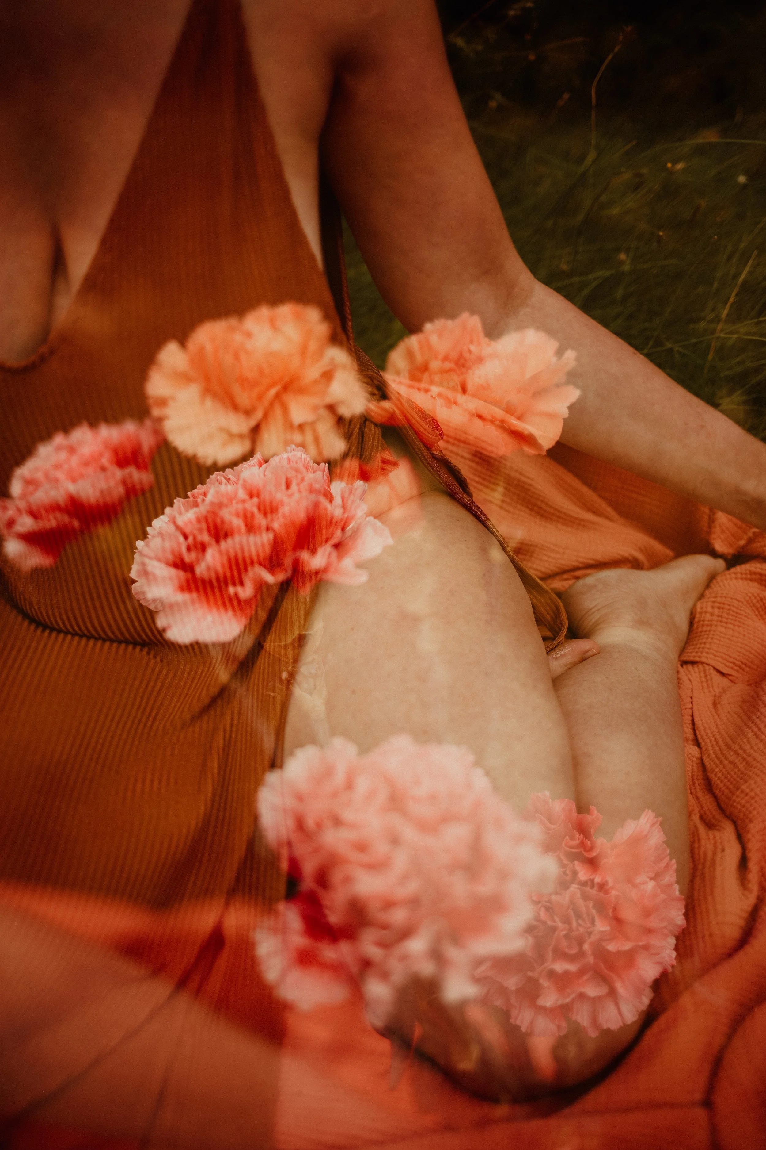 Une personne assise sur de l'herbe, portant une robe orange avec des fleurs en tissu ou en broderie dans des tons roses, avec des fleurs roses en décoration ou accessoires.
