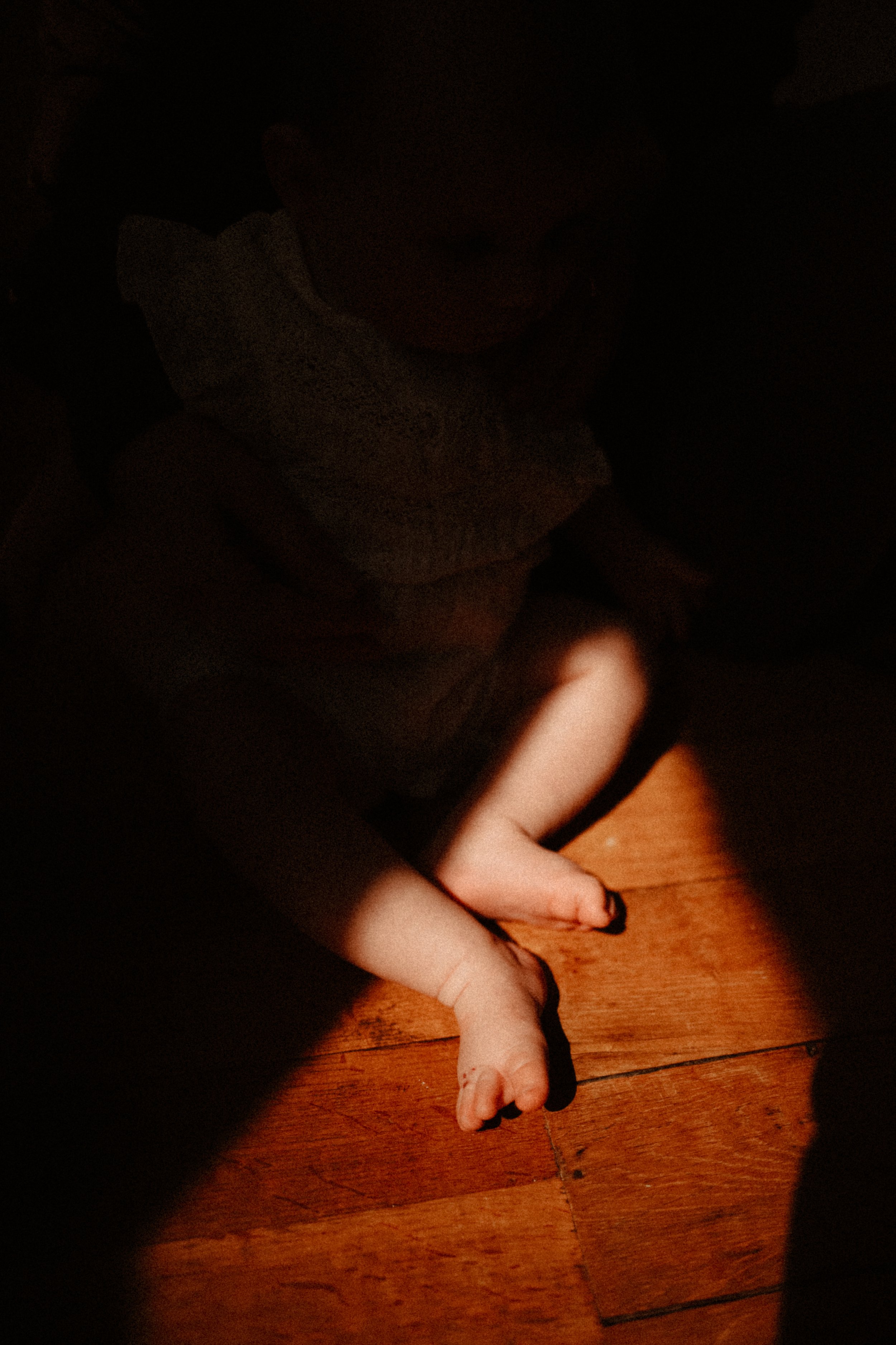 Un enfant assis sur un plancher en bois, vêtu d'un t-shirt gris et portant des chaussettes noires, dans une pièce sombre.